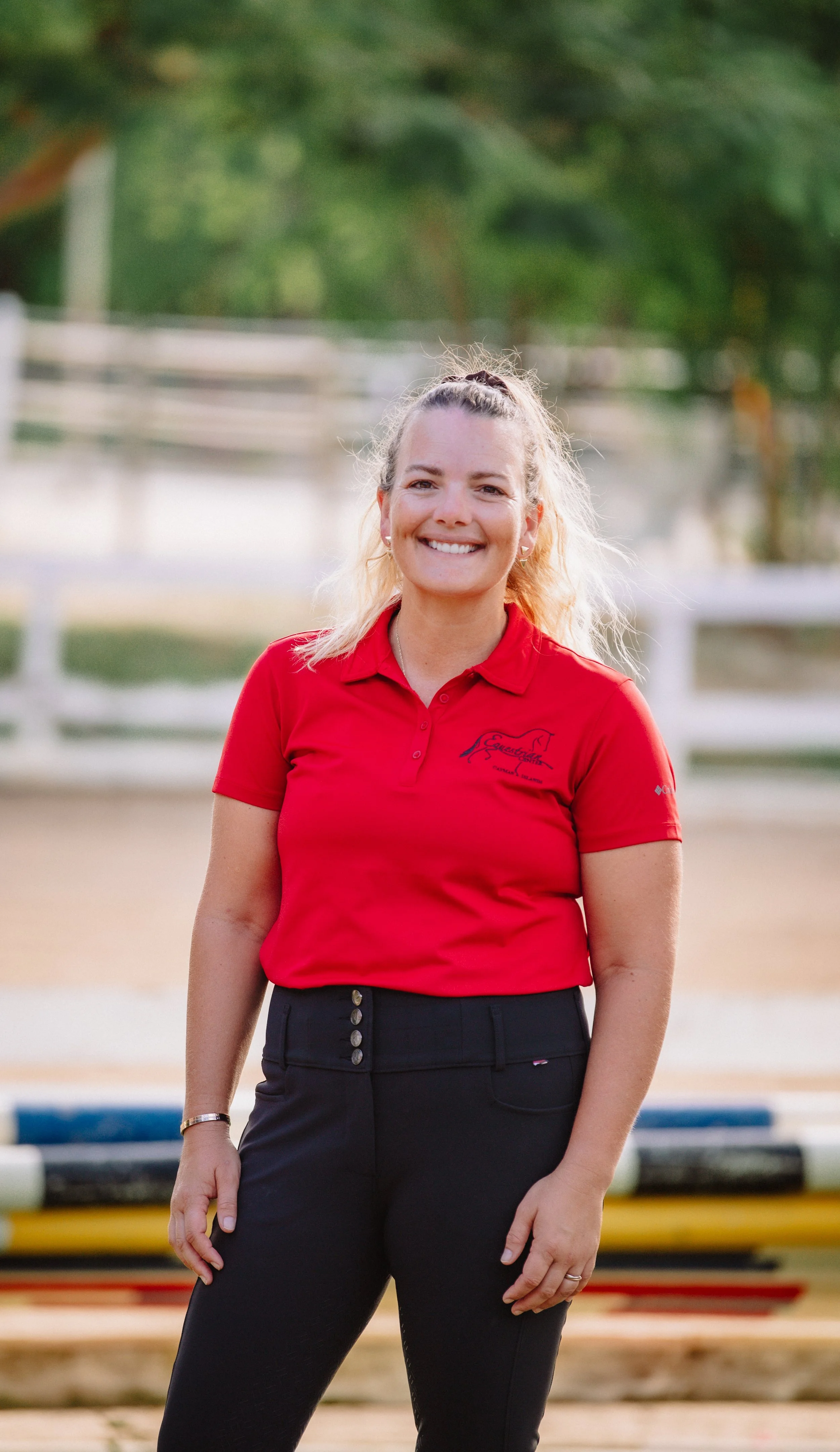 A woman with blonde hair tied in a ponytail, wearing a red polo shirt and black pants, standing outdoors near a rowing boat and a dock, smiling at the camera.