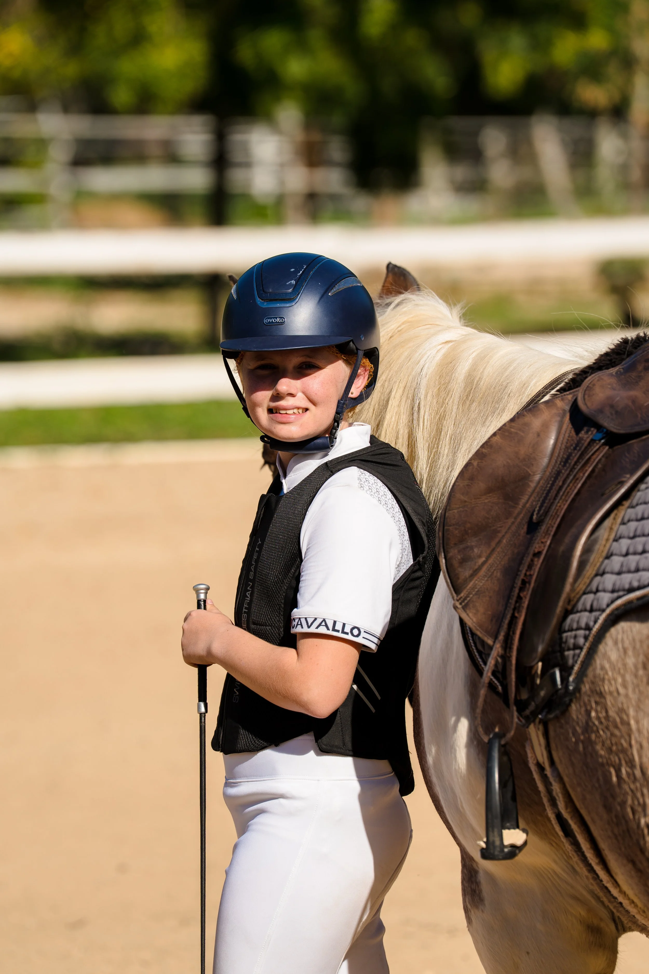 A young female equestrian standing outdoors with a white horse, wearing a black riding helmet, white riding breeches, and holding a riding crop.