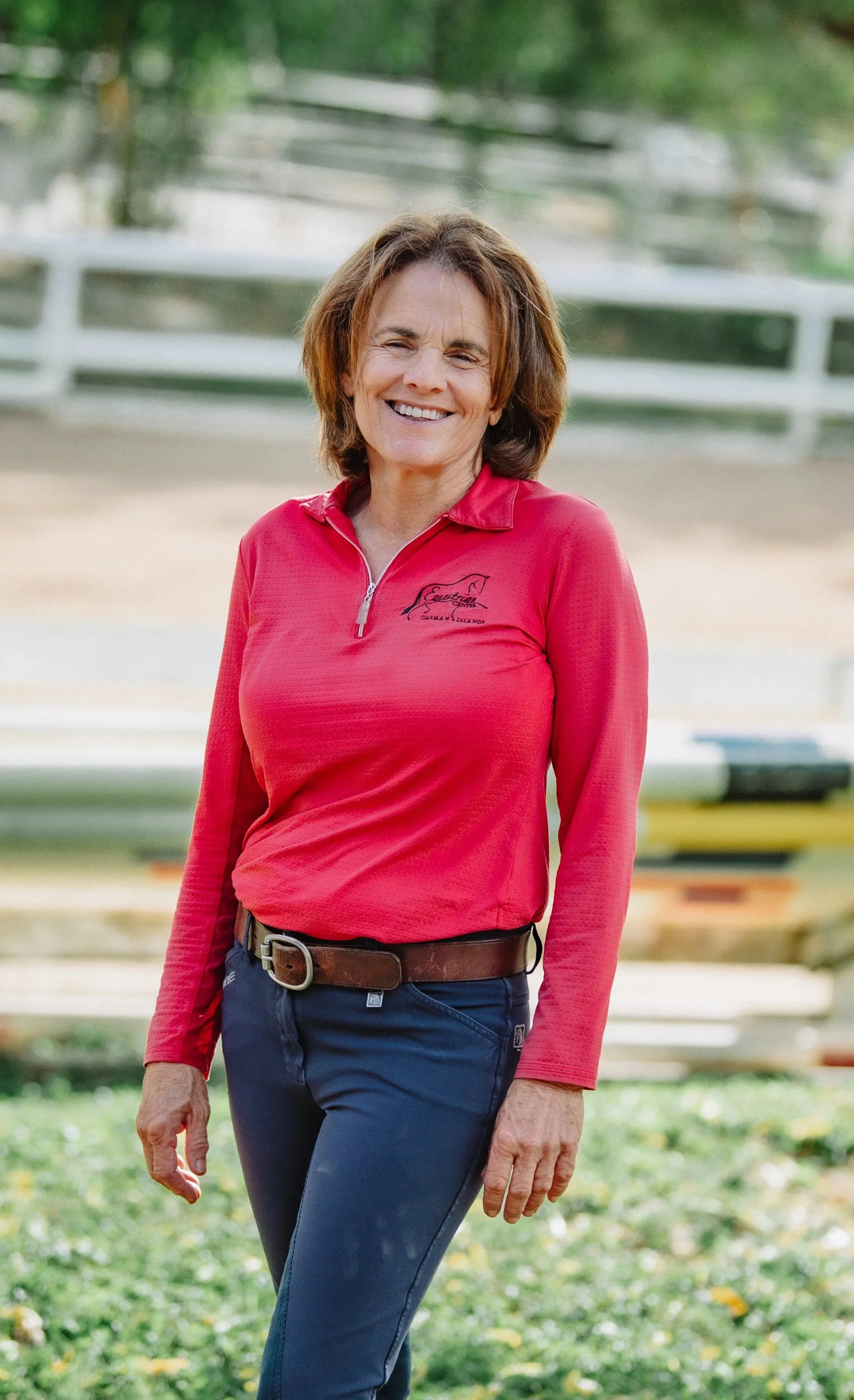 A middle-aged woman with short brown hair smiling outdoors in front of a white fence, wearing a red zip-up shirt with a logo, black pants, and a brown belt.
