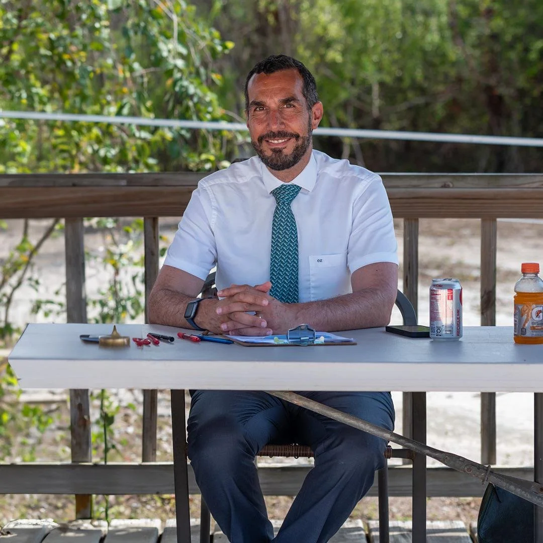 A man in a white shirt and teal patterned tie sitting at a table outdoors. On the table are a clipboard, red and black pens, a can of soda, an orange Gatorade, and a clipboard. The man is smiling and has his hands clasped in front of him. There are trees and a wooden railing in the background.