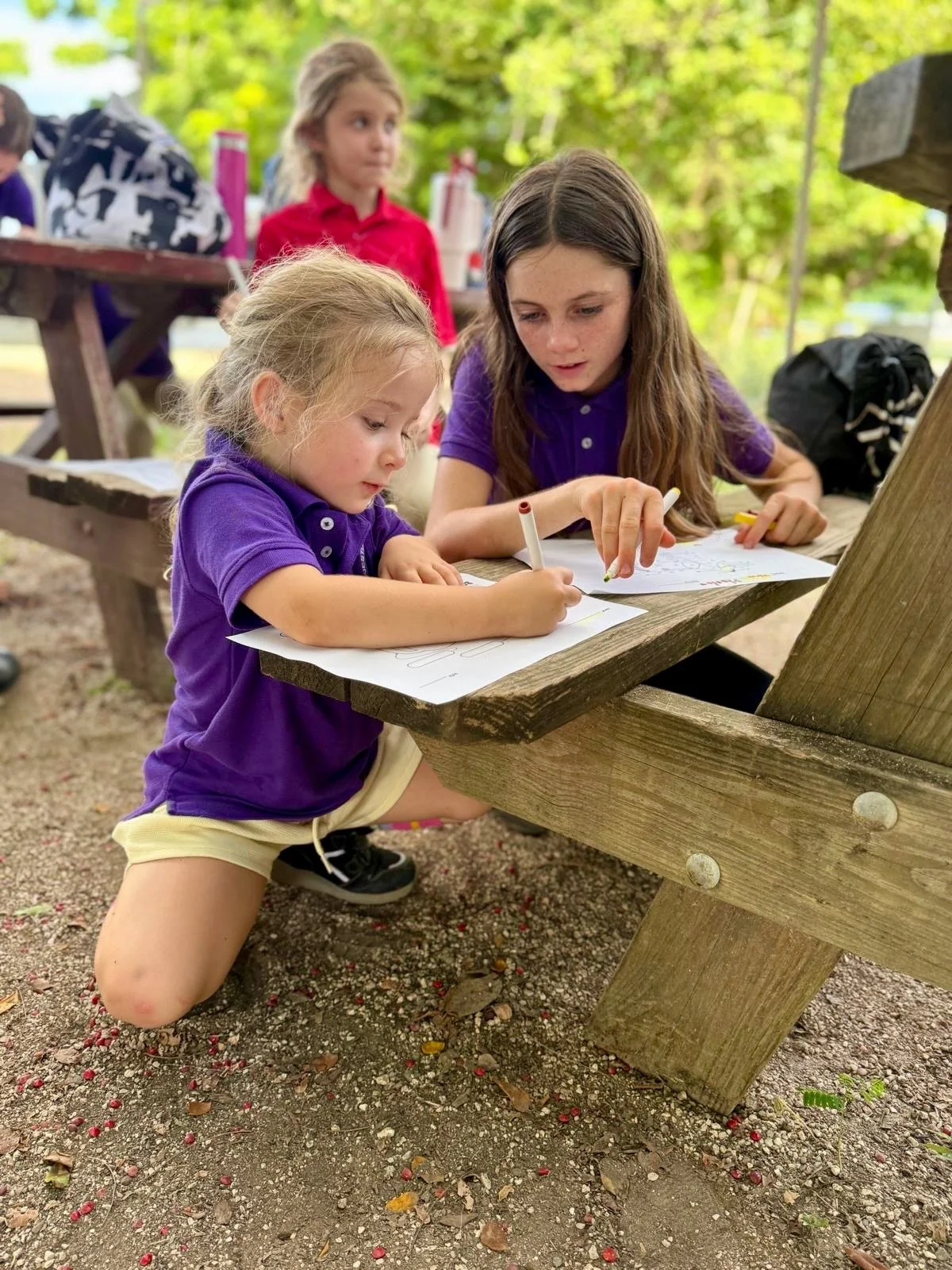 Two young girls are drawing at a wooden outdoor picnic table with papers and markers. One girl with long brown hair in a purple shirt is helping a younger girl with curly blonde hair in a purple shirt and yellow shorts with her drawing. They are surrounded by a natural outdoor setting with trees.