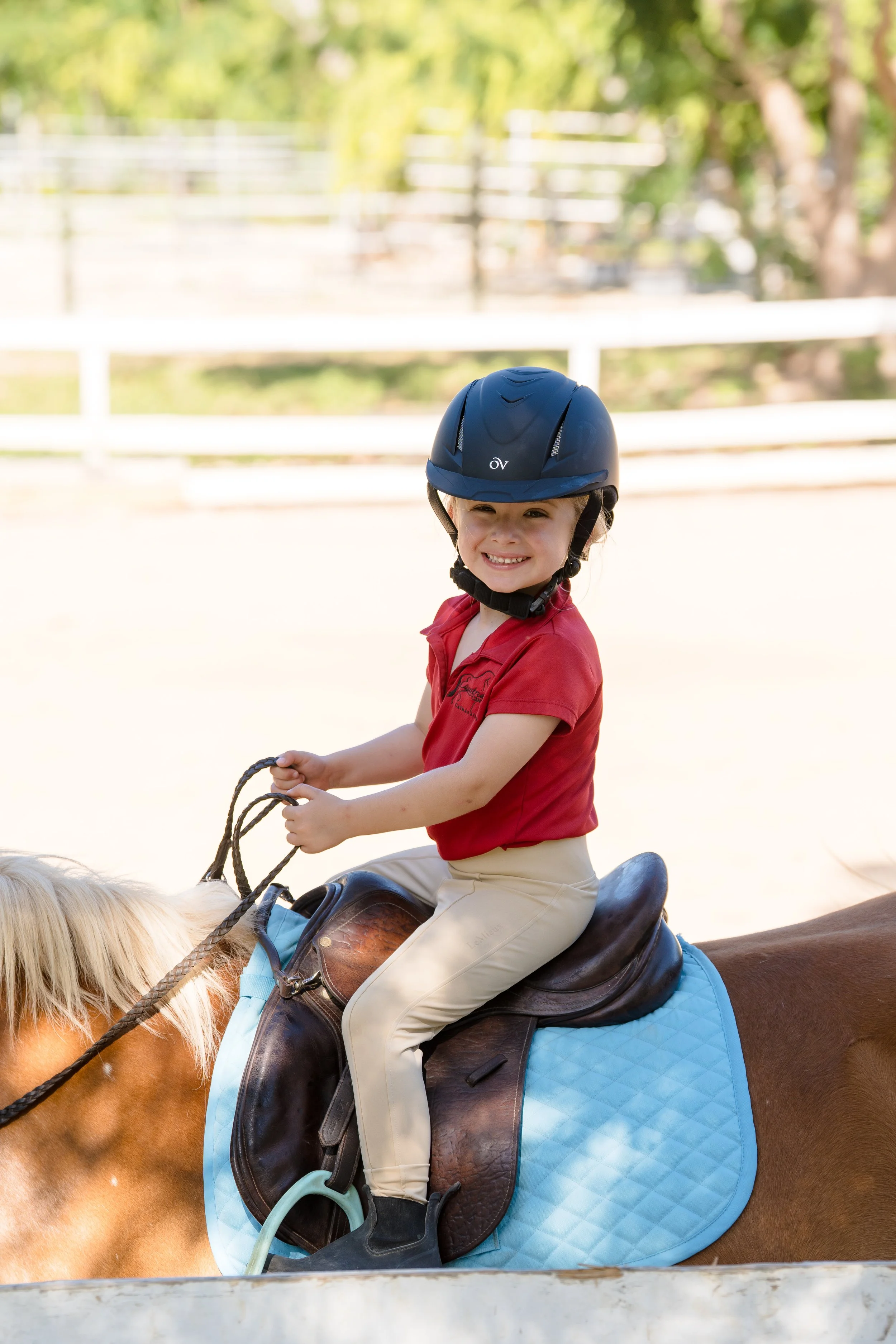 A young girl with a helmet riding a horse, smiling, outdoors on a bright day.
