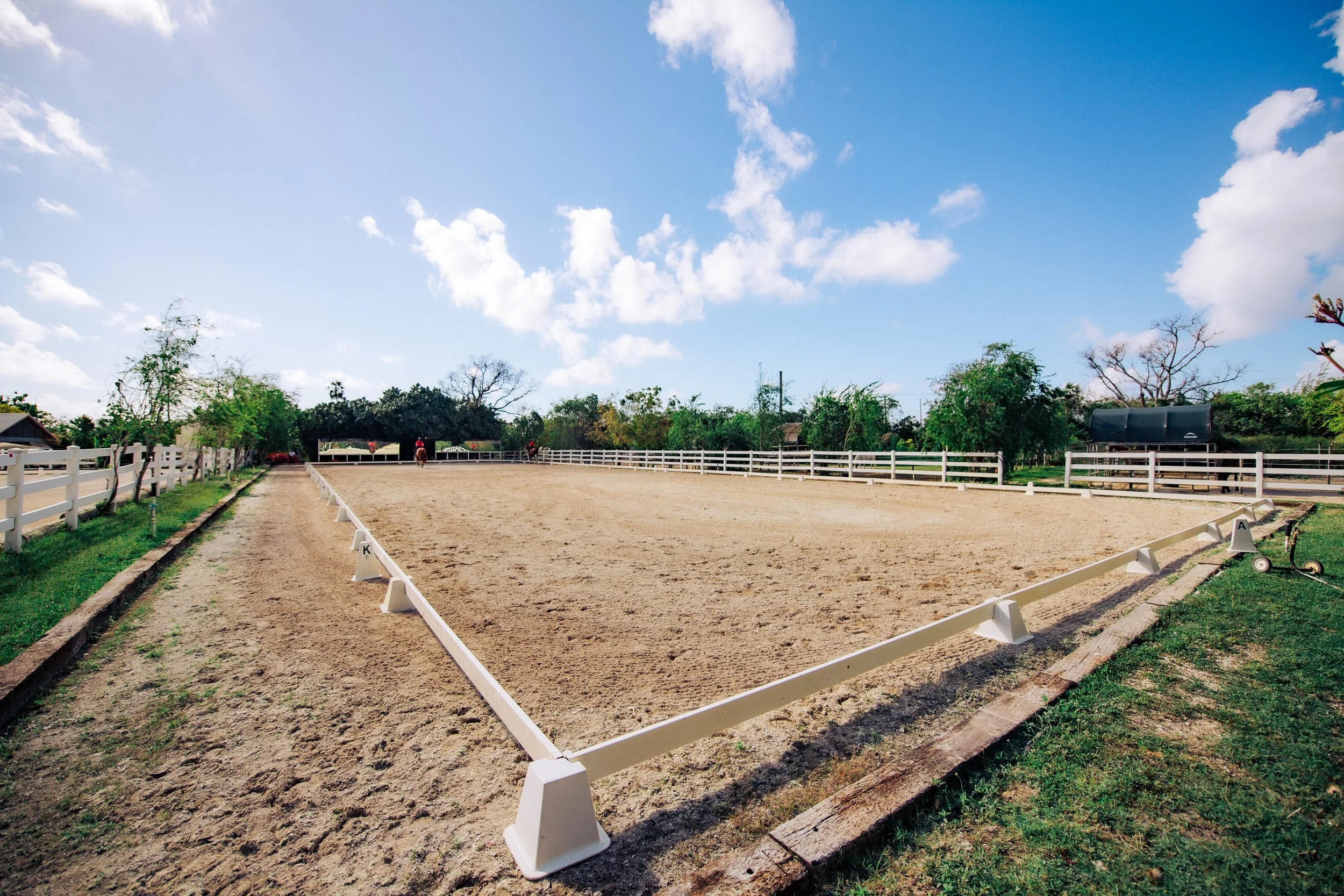 Equestrian riding arena with a sandy surface, white rail fencing, and riders on horseback under a partly cloudy blue sky.