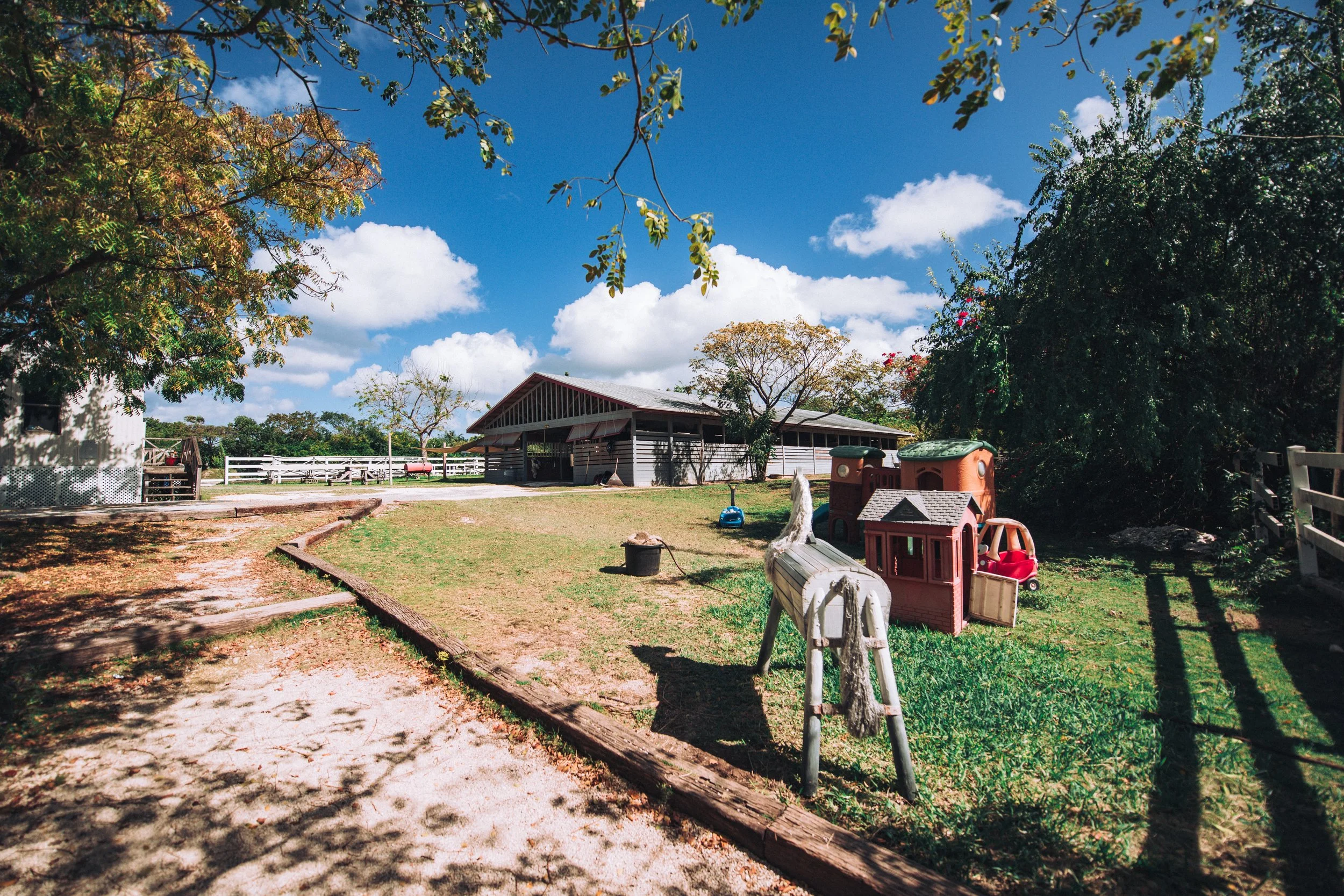 A fenced outdoor play area with small houses, toys, and trees under a clear blue sky.