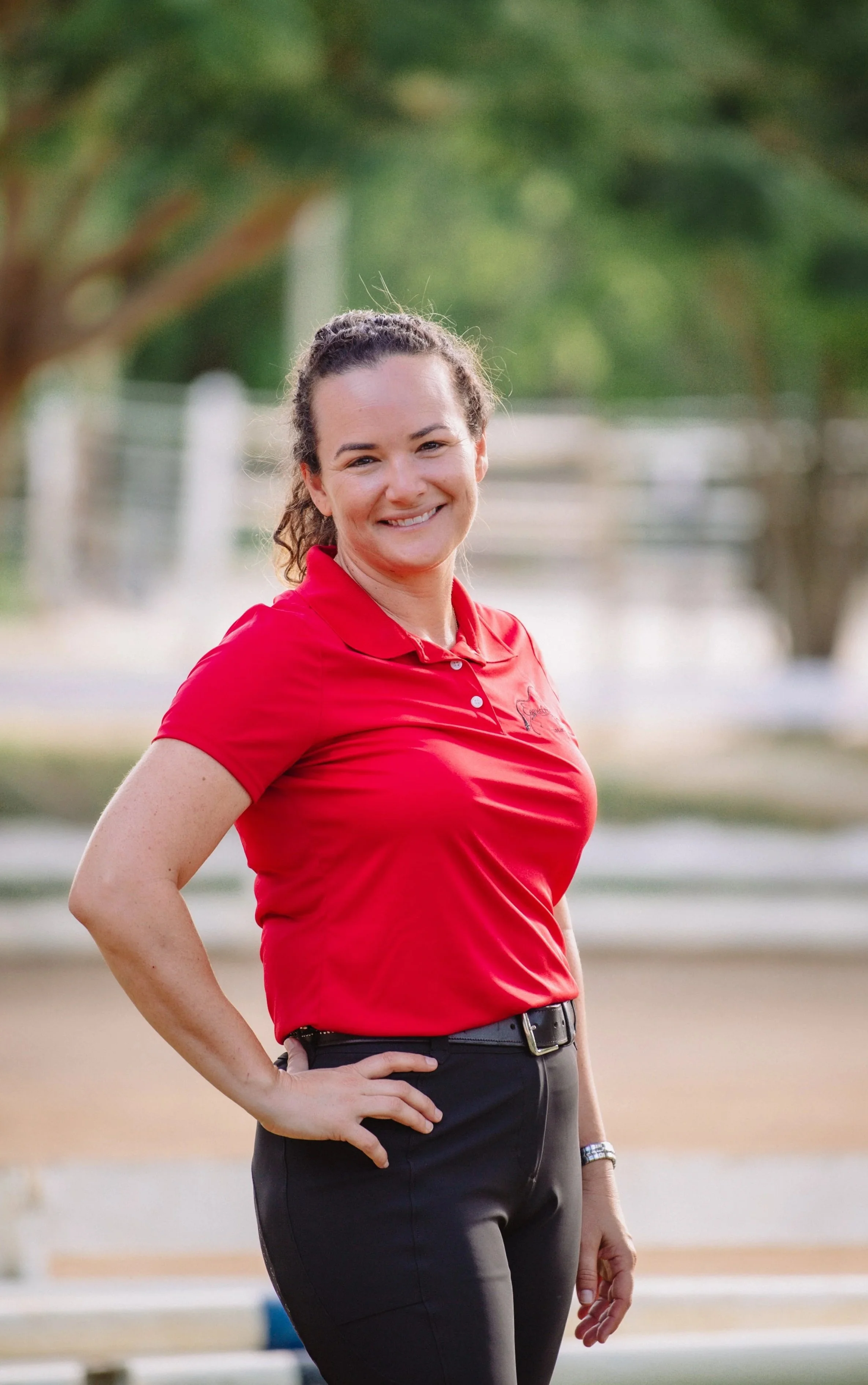 A woman in a red polo shirt and black pants standing outdoors with her hand on her hip, smiling at the camera.