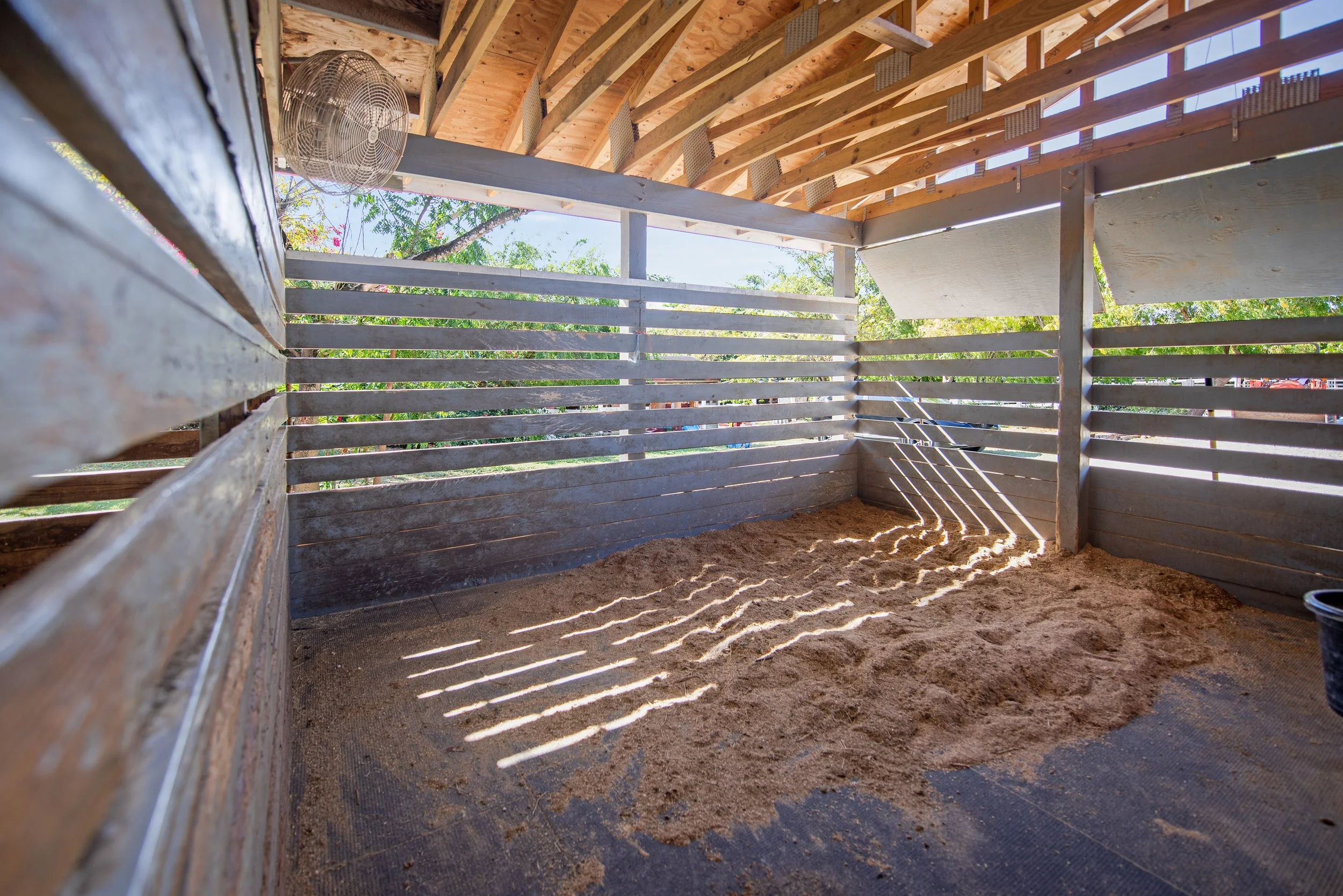 Inside view of a wooden horse stable with partially built walls and a roof, shadow patterns on the ground, and a fan mounted on the wall.