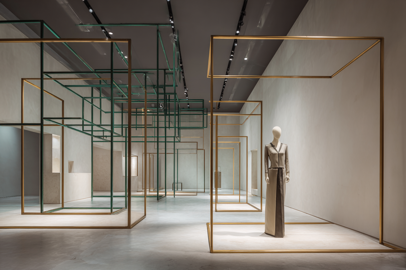 Empty fashion retail display with a mannequin dressed in gray and beige clothing, surrounded by geometric gold and green metal frames in a modern, minimalist store interior.