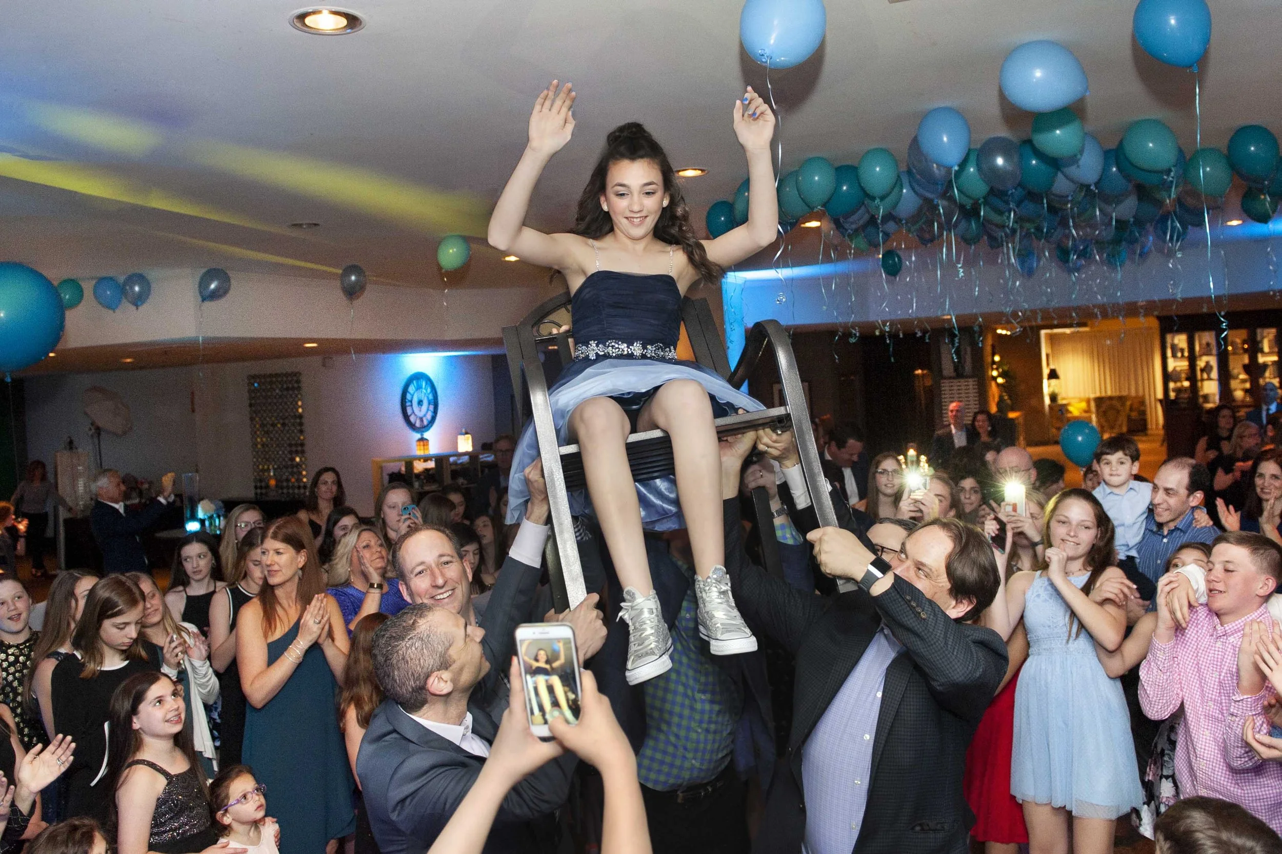 A girl in a blue dress sits on a chair being lifted by several adults at a celebration with balloons and many people taking photos and clapping.