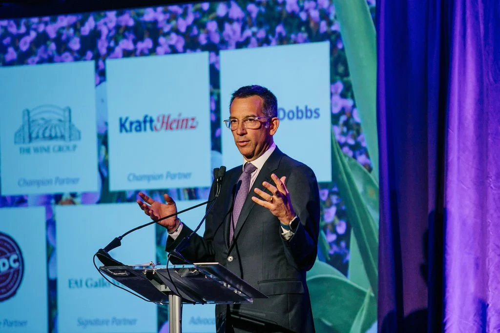 A man in a suit and glasses is speaking at a podium during a presentation or conference with a purple and green backdrop featuring sponsor logos.