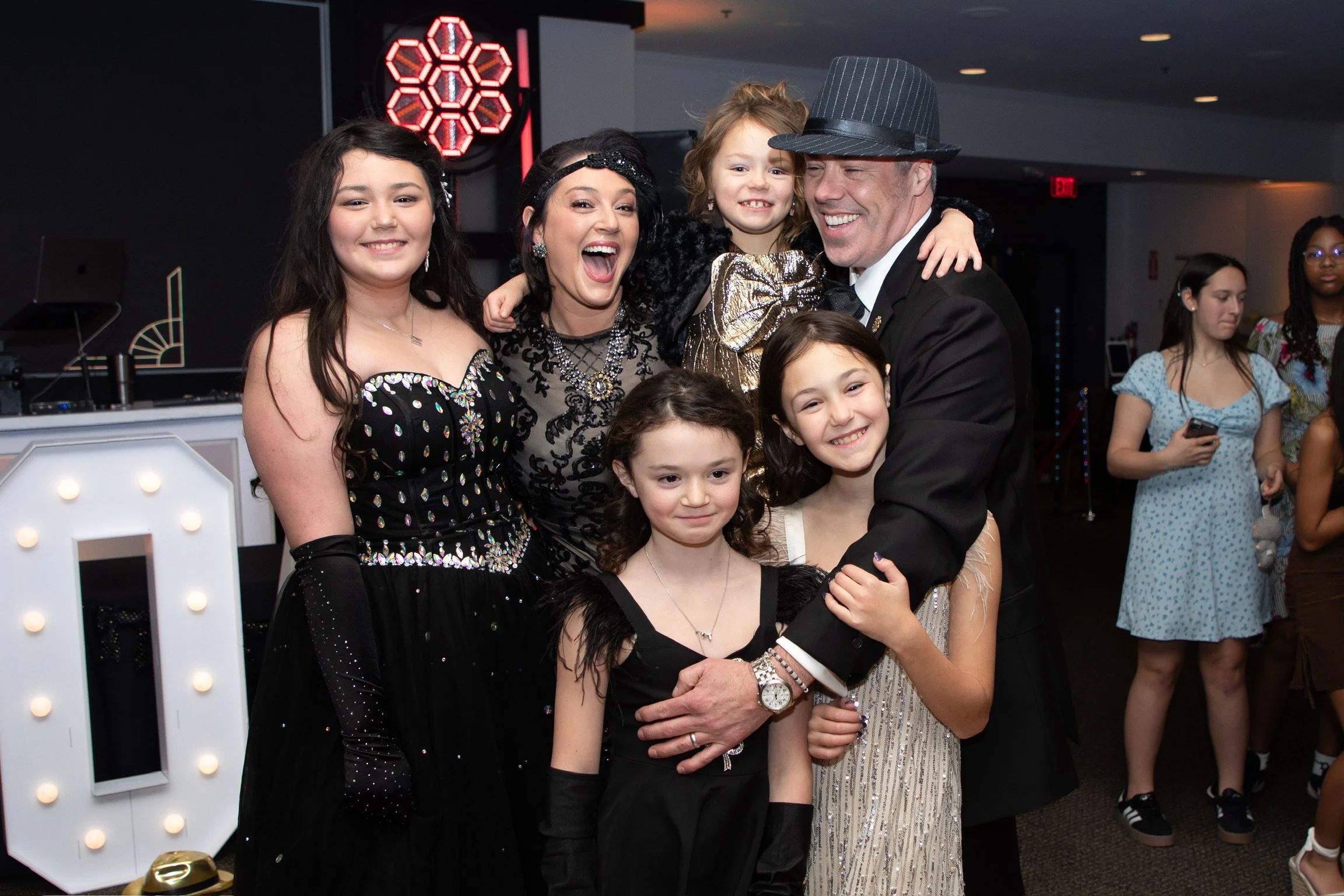 A group of seven people, including children and adults, smiling and hugging at a celebration or party in an indoor venue.