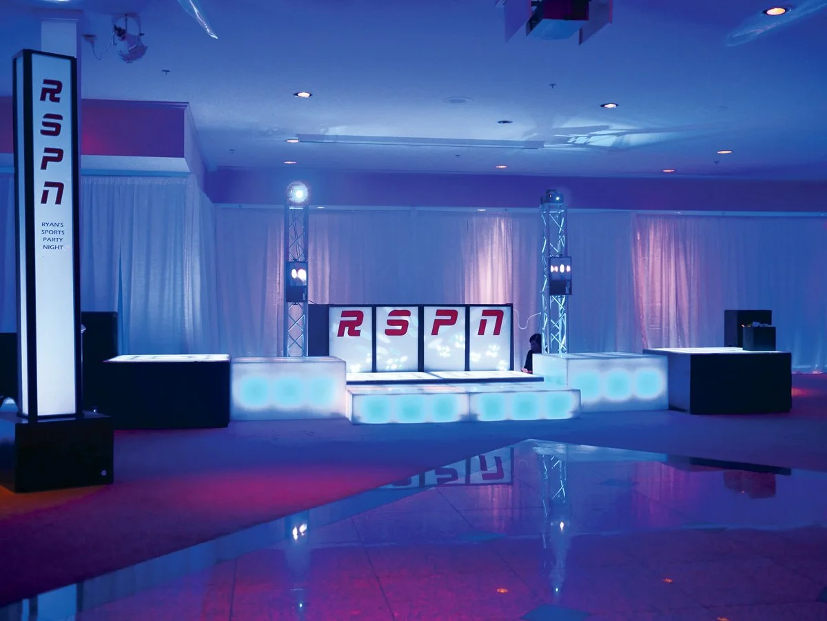 An empty event stage with lighting and DJ equipment, featuring a backdrop with the letters R, S, P, N, and a banner on the side reading RYAN'S SPORTS PARTY NIGHT.