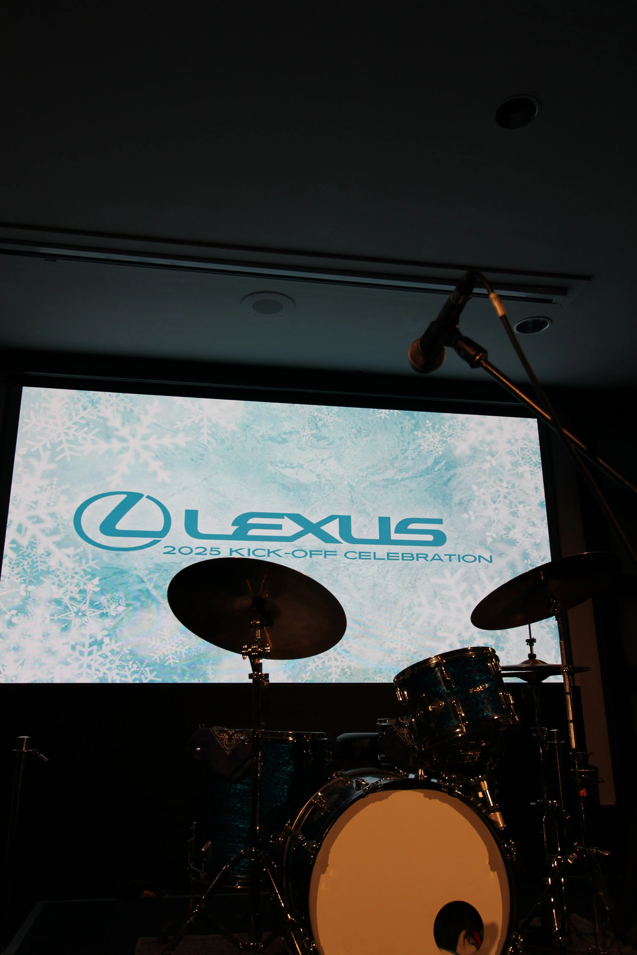 Stage with a drum set in front of a large screen displaying the Lexus logo and the text "2025 Kick-Off Celebration" in front of a snowy background.