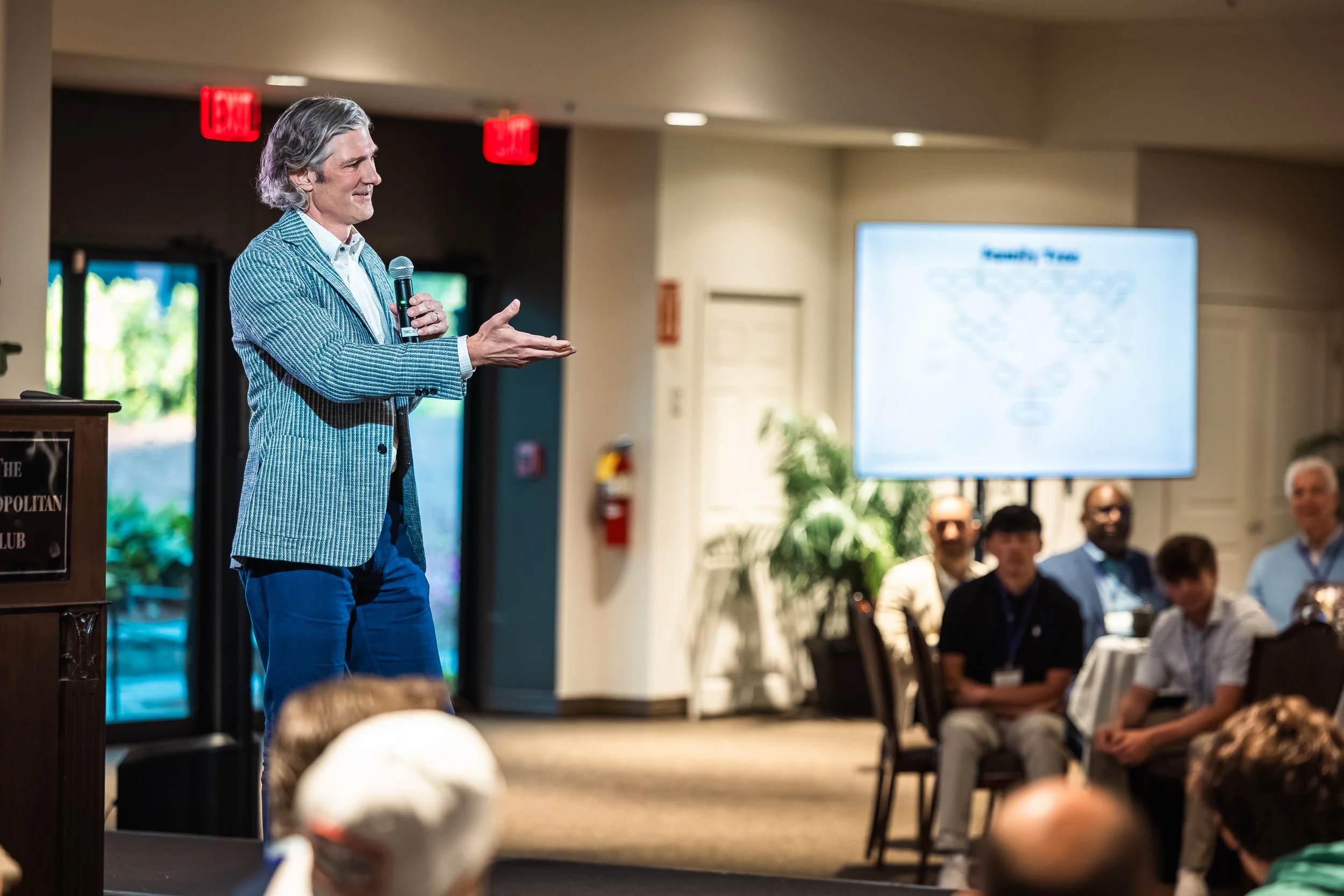 A man in a blue blazer and blue jeans holding a microphone, speaking to an audience in a conference room with a presentation screen and seated attendees.
