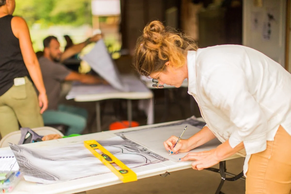 A woman wearing glasses and a white shirt is leaning over a table, drawing or marking on a large sheet of paper with a pencil. There is a yellow level tool on the paper. In the background, other people are working on different projects in a bright in