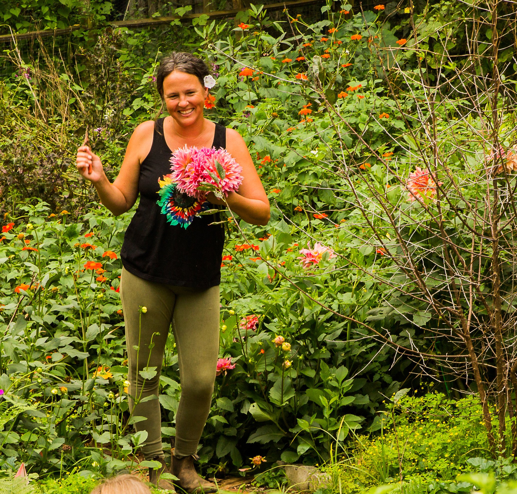 A woman smiling, holding a bunch of colorful flowers in a garden with green foliage and vibrant orange and pink flowers.