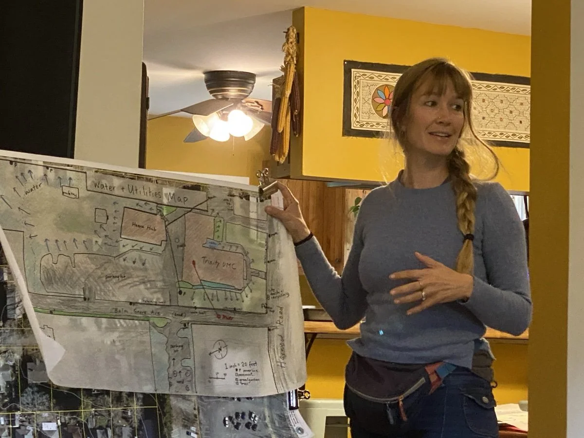 A woman with a braid in a gray long-sleeve shirt stands beside a detailed hand-drawn map of water and utilities, seen indoors with a ceiling fan and yellow walls in the background.