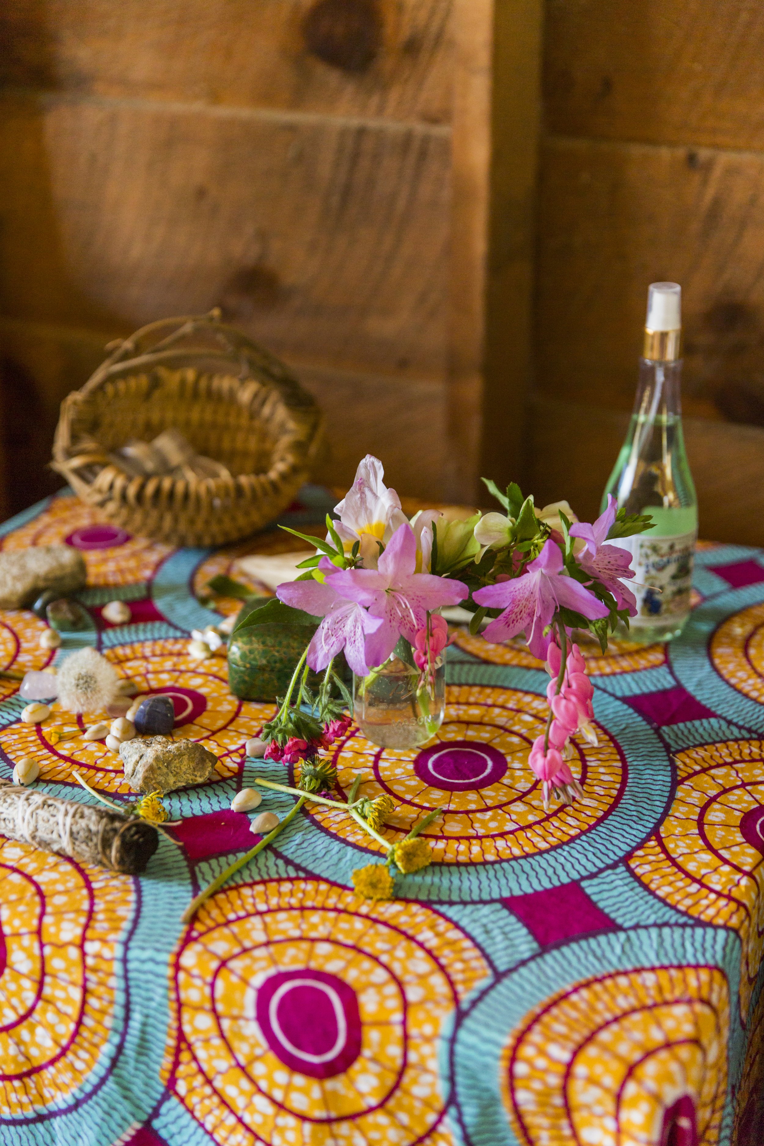 A table covered with a vibrant, colorful cloth displaying a bouquet of pink and white flowers in a small glass vase, along with various small objects including stones, shells, a rolled item, and a spray bottle, against a wooden background.