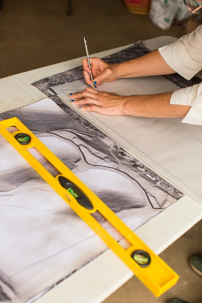 Person marking a landscape plan with a pen on a table, with a yellow level tool nearby.