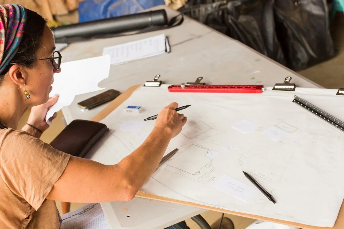 A woman sitting at a table drawing on large paper with various drafting tools around her