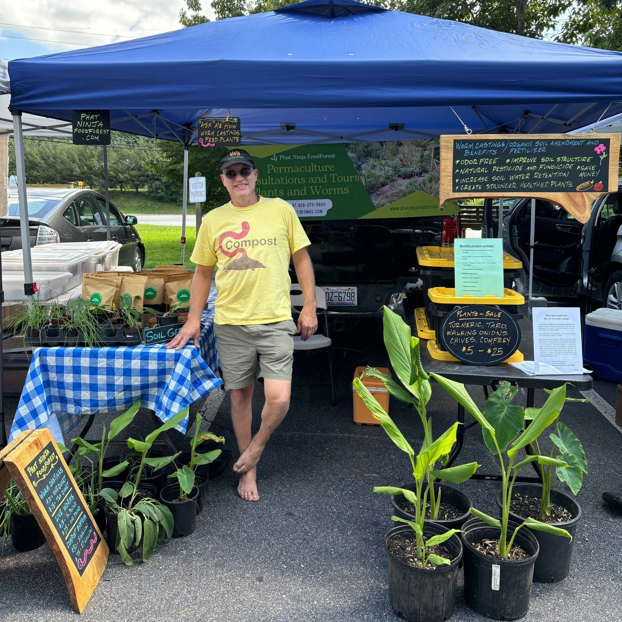 A man wearing sunglasses, a yellow t-shirt with a compost logo, and shorts, standing barefoot at a plant sale booth under a blue canopy. The booth features potted plants and handwritten signs about worm castings and soil amendments, with cars in the background and green trees.