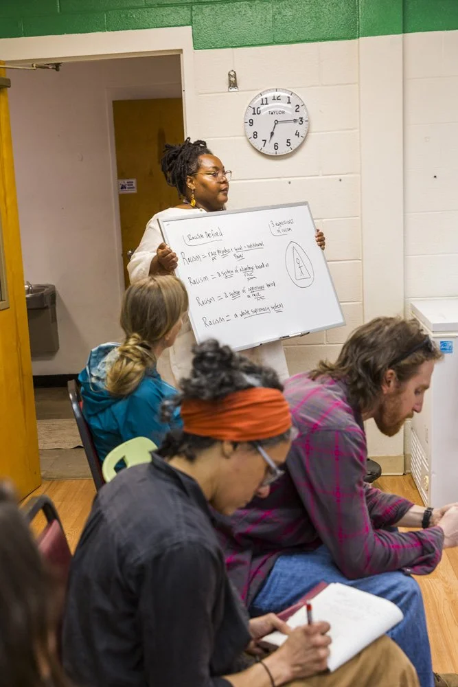 A woman with glasses and earrings stands at the front of a classroom holding a whiteboard with notes and diagrams. Four students sit listening, taking notes, and looking at her. A clock on the wall shows 12:37, and the room has green and white painted walls.