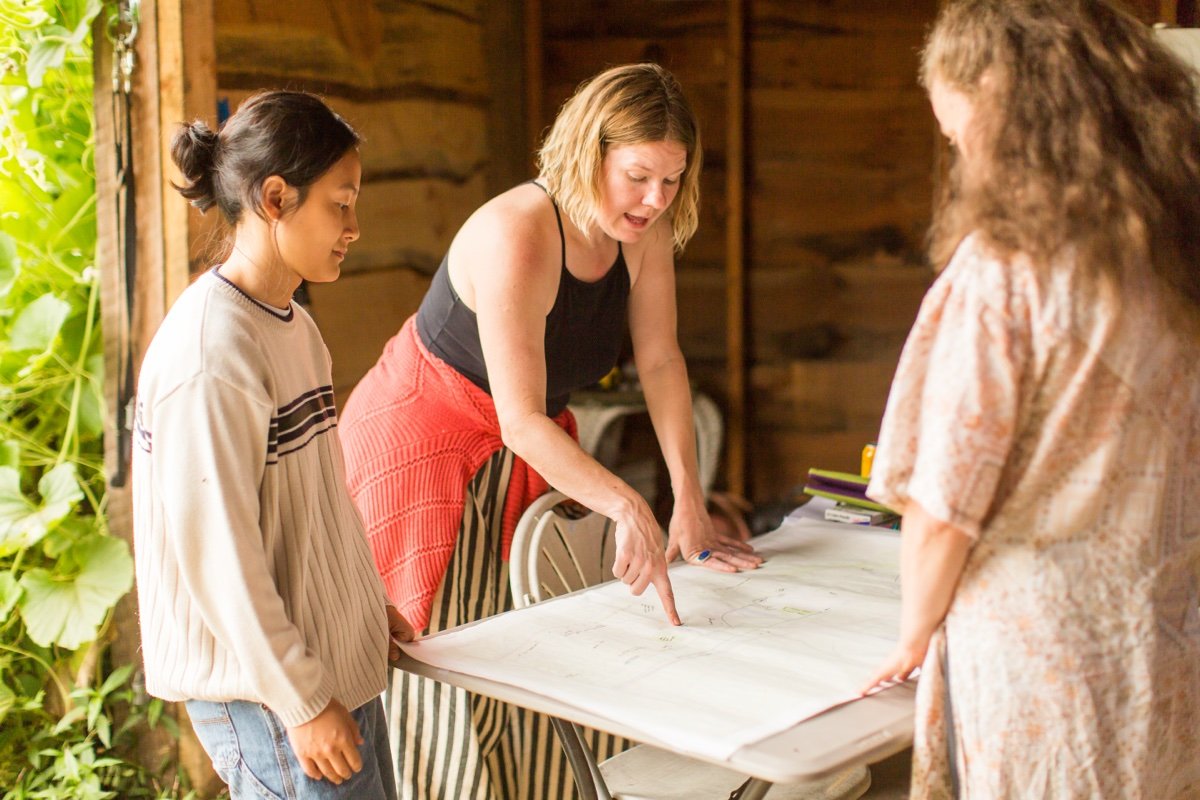 Three women gathered around a table in a rustic, wood-paneled room, examining a large sheet of paper or blueprint.