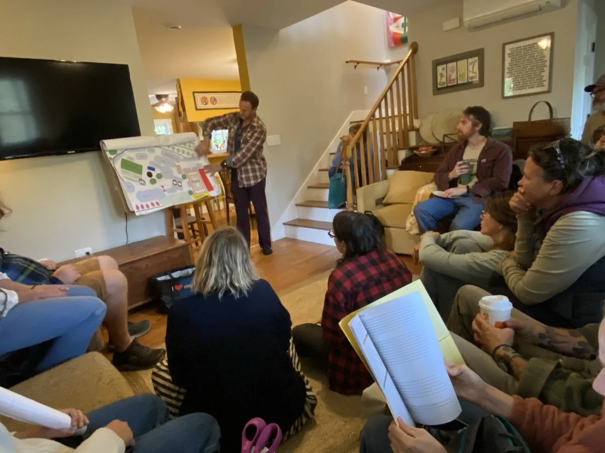 A group of people attending a presentation in a cozy living room, with a man standing and pointing at a large colorful map or diagram on an easel. The audience sits on sofas, chairs, and the floor, listening attentively, some holding notebooks, drink