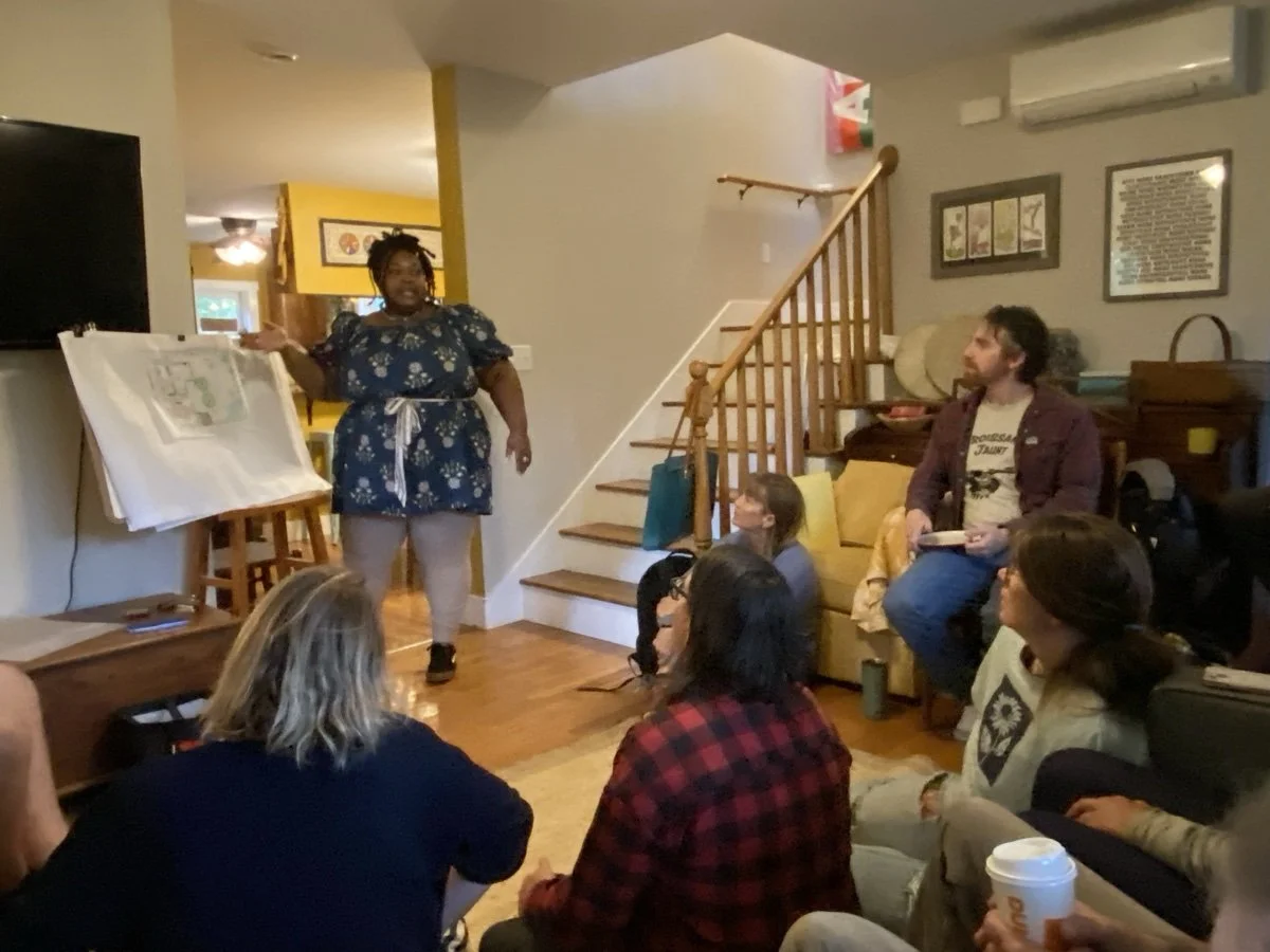 A woman standing near a whiteboard giving a presentation or storytelling to a seated audience in a home setting. Several people are seated on the floor and on a couch, listening attentively.