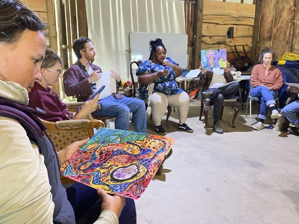 Group of people sitting in a room with wooden walls, some holding artwork and looking at each other, participating in an art or discussion session.