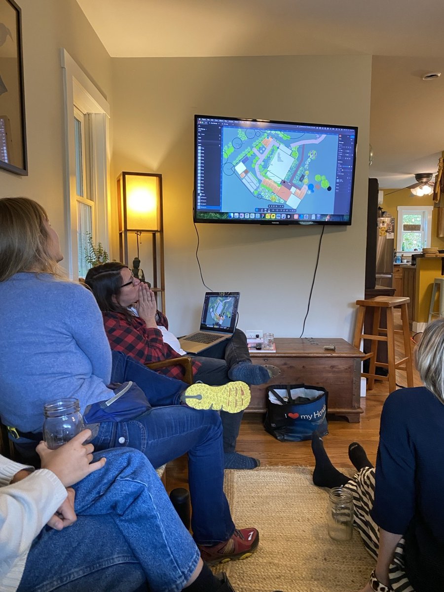People gathered in a living room watching a large screen displaying a design or plan, possibly for landscaping or construction.