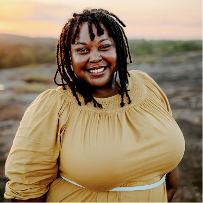 A woman smiling outdoors during sunset, wearing a yellow dress and standing on rocky terrain.