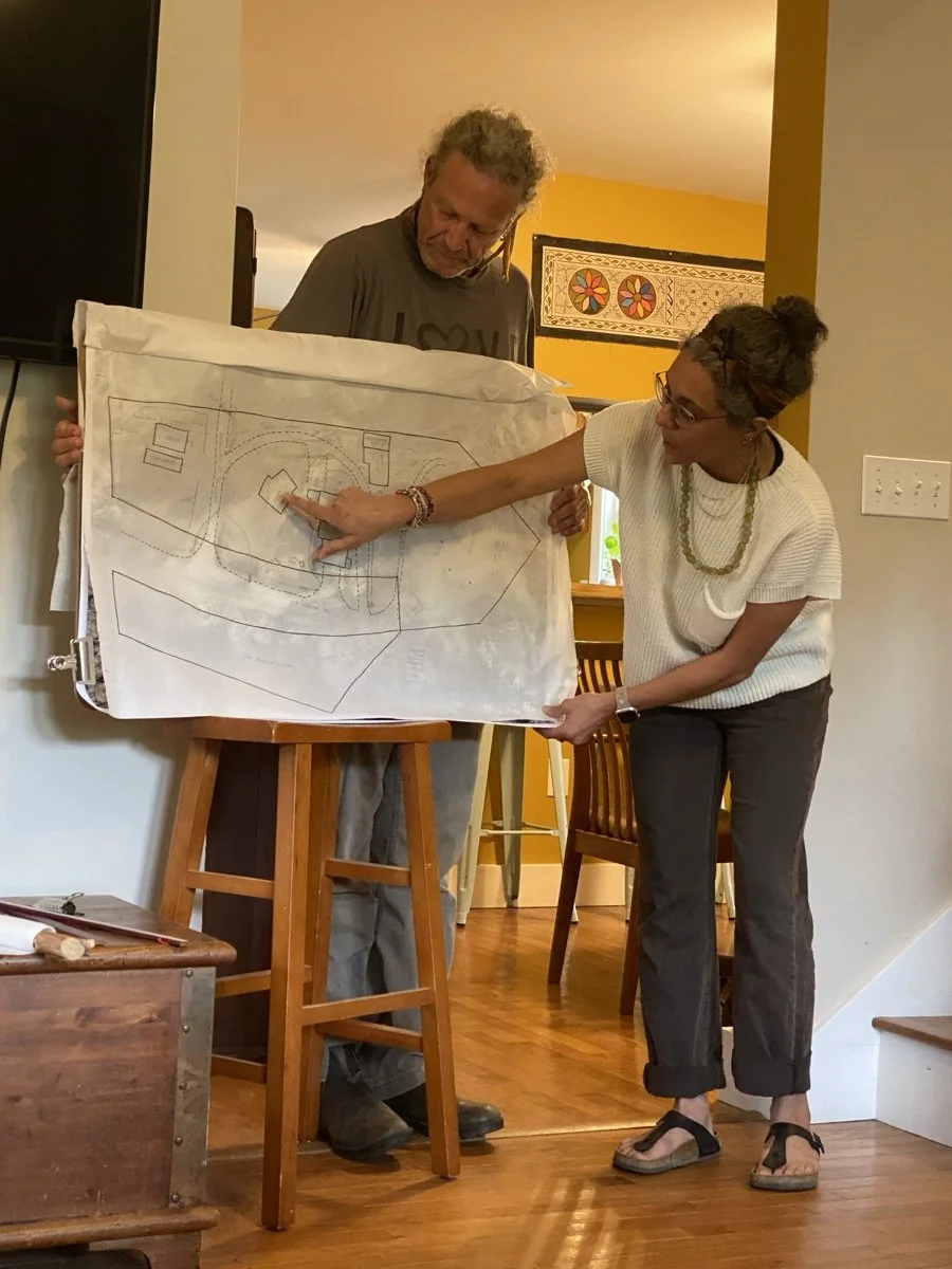 Two people, a man and a woman, are looking at and pointing to a large architectural blueprint or site plan on an easel in a home setting.