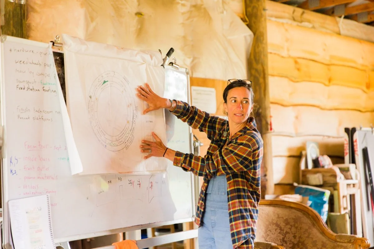A woman in a plaid shirt is giving a presentation about composting or soil health indoors, with diagrams on a whiteboard.