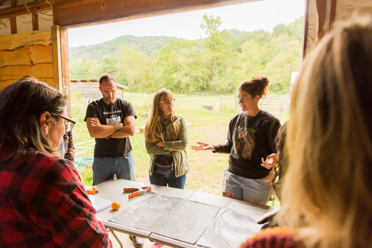 A group of people gathered around a table in a wooden shed, listening to a woman with glasses and a black sweatshirt with a tiger graphic, who is speaking and gesturing with her hands. Outside, there is a green landscape with trees and hills.