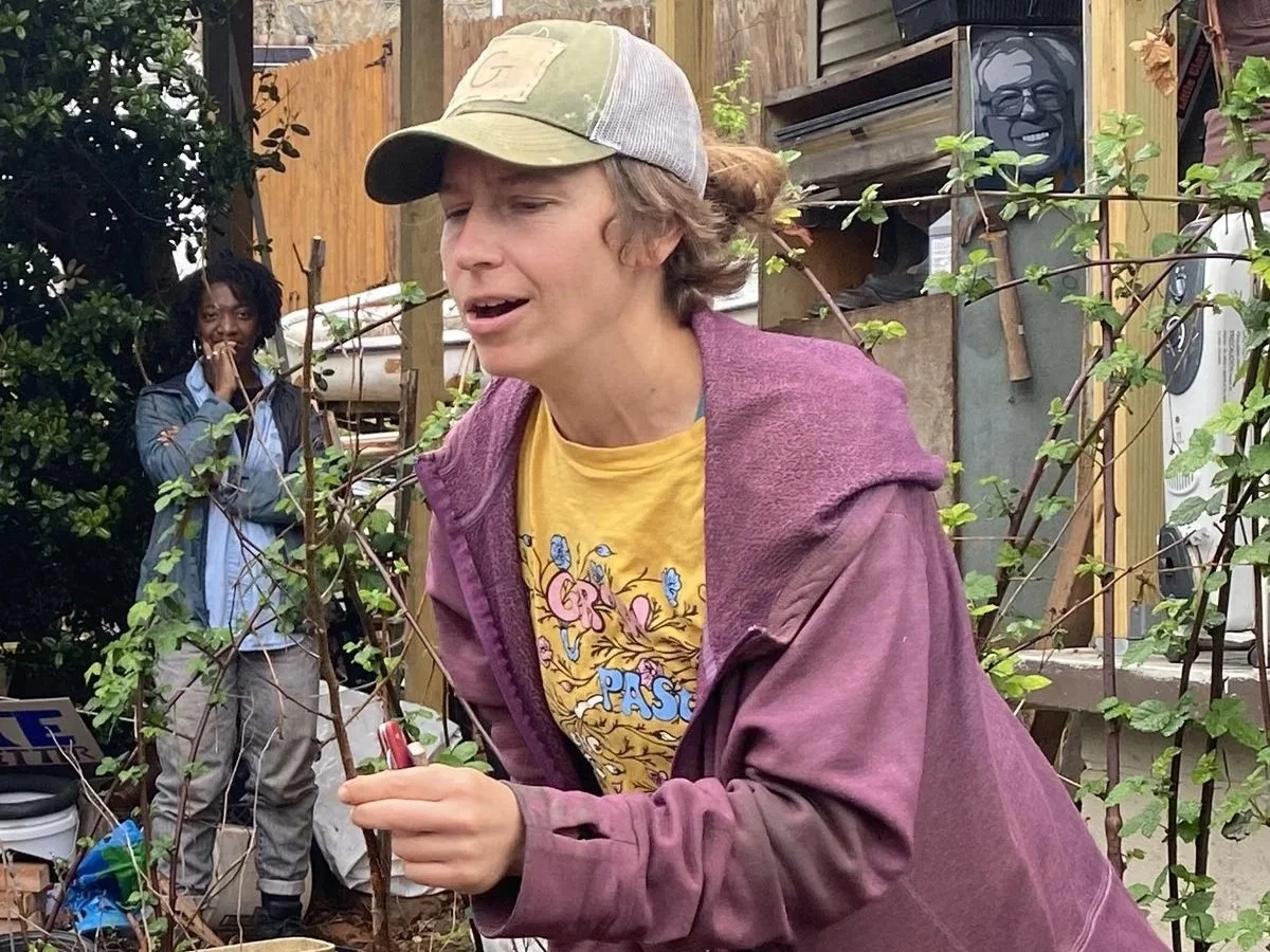 A young person with shoulder-length brown hair, wearing a beige and gray cap, purple hoodie, and yellow graphic T-shirt, bending forward outdoors among plants and trees, holding a small object, with a surprised or engaged facial expression. In the background, a woman with braided hair, wearing a denim jacket over a blue shirt, stands with her hand near her face. There are wooden structures, plants, and outdoor items visible in the setting.