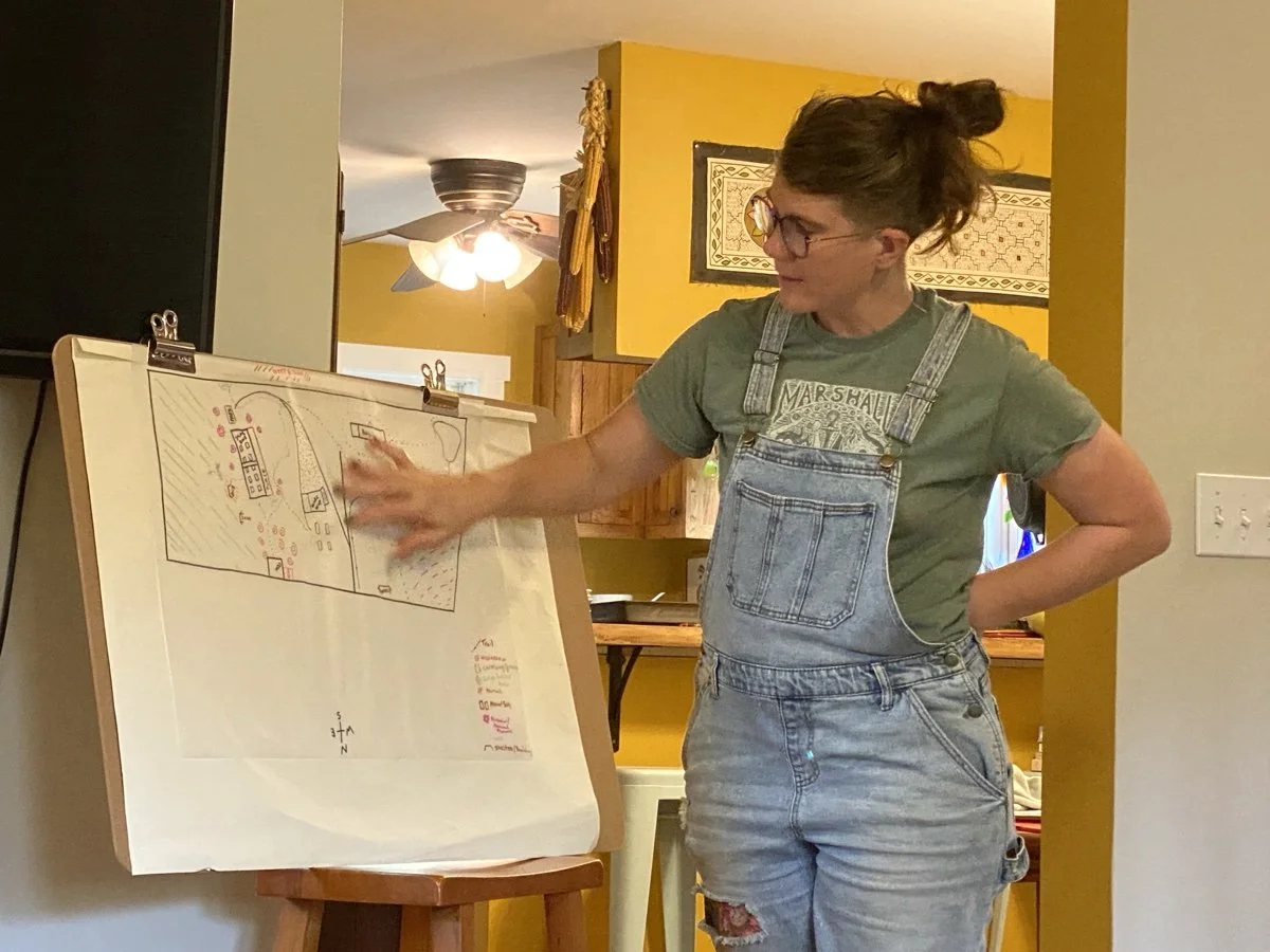 A woman with glasses and brown hair tied in a bun wearing a green t-shirt and denim overalls pointing to a large paper map on a stand.