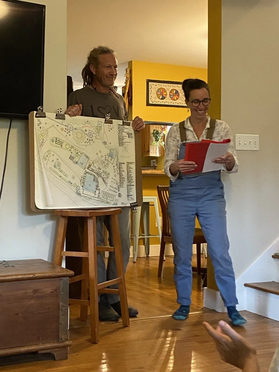 Two people standing in a room, one holding a large layout map and the other reading from a notebook, with a person reaching out in the foreground.