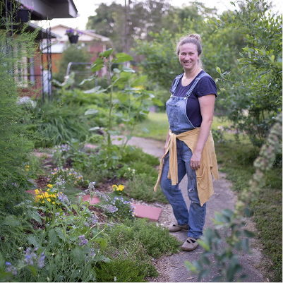 A woman standing on a garden path next to flowering plants and greenery, smiling at the camera.