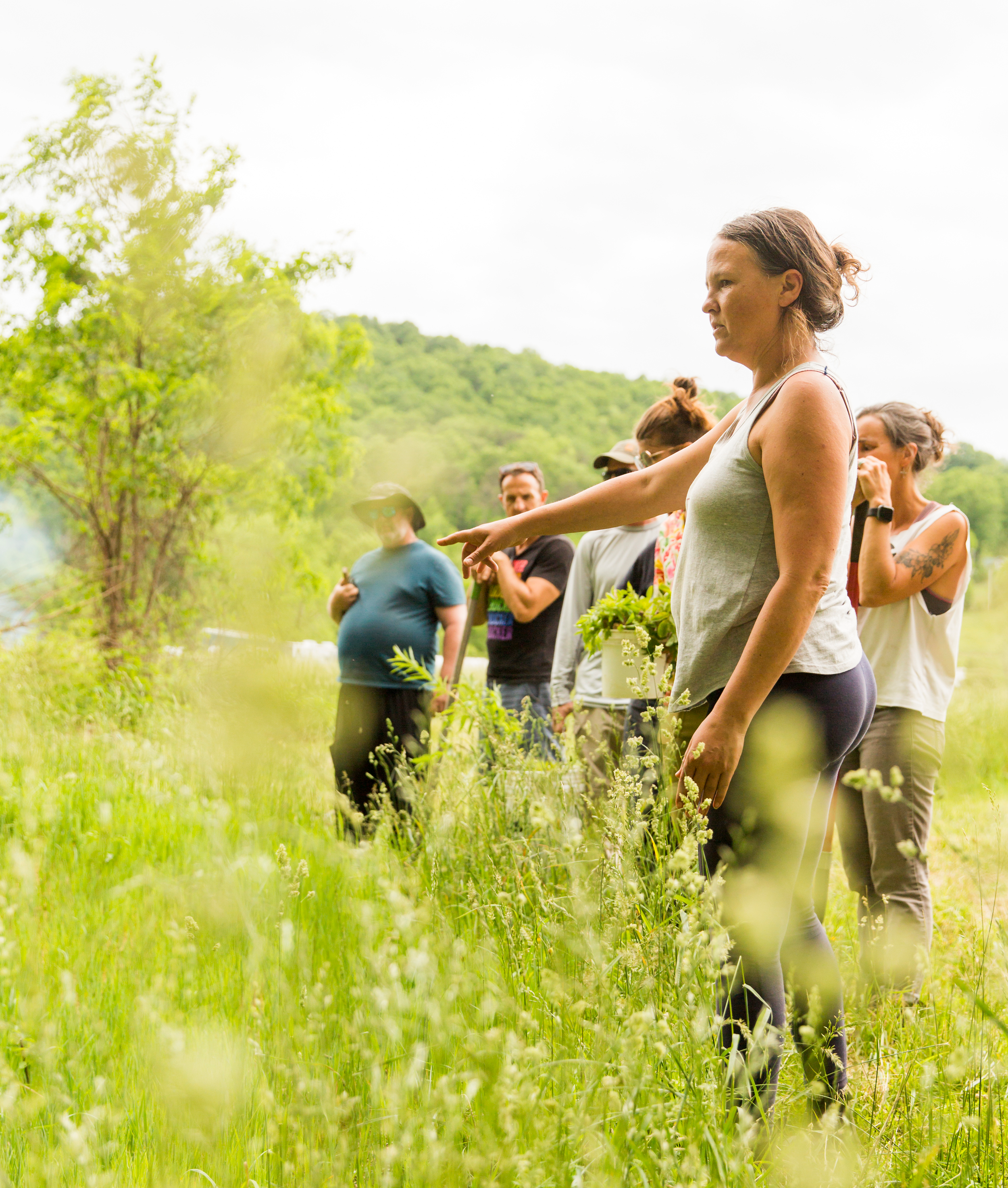 A woman in a gray tank top pointing towards plants in a green outdoor setting, with a group of people standing behind her and a scenic landscape of trees and hills in the background.