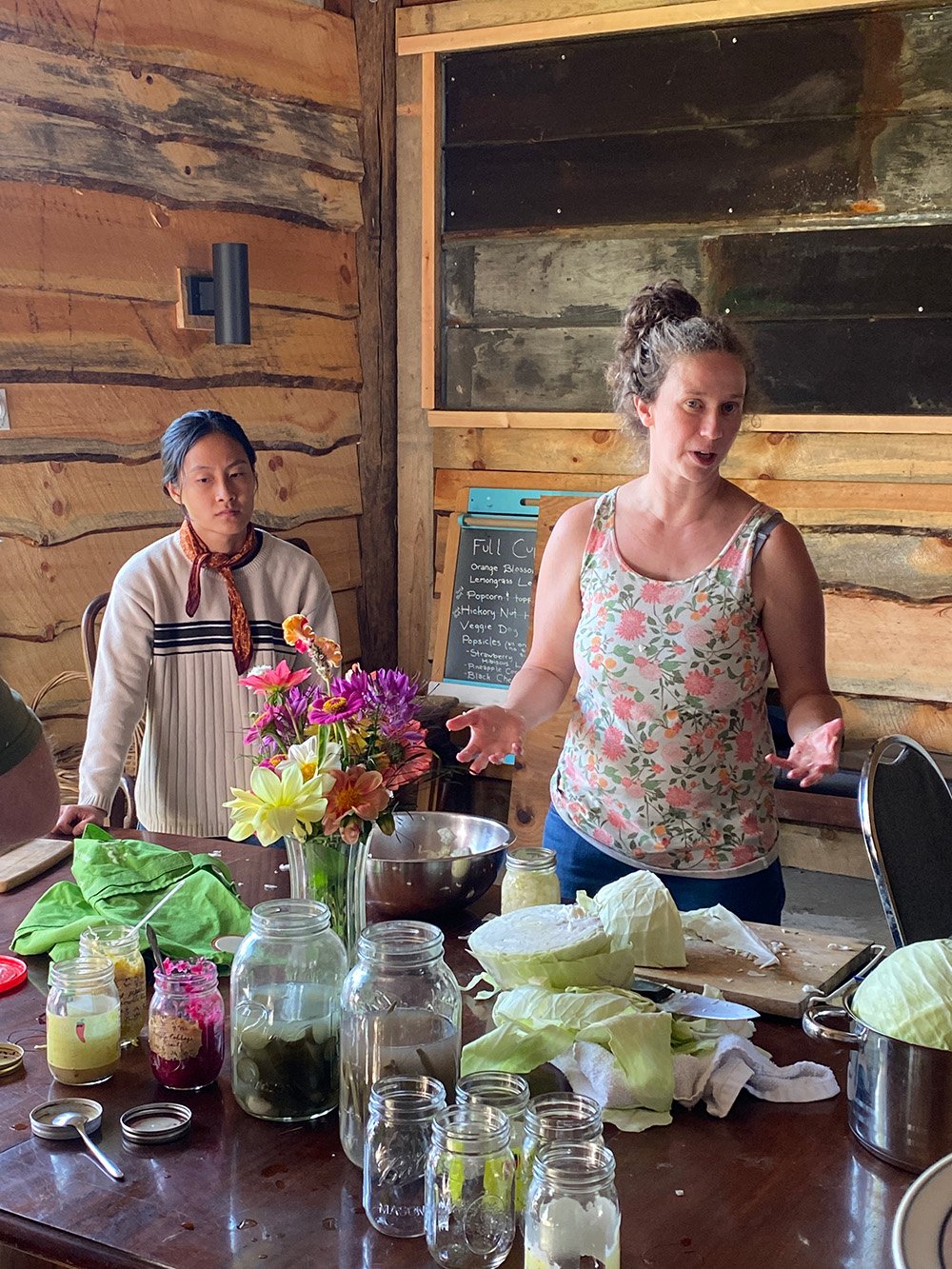 A woman in a floral sleeveless top gestures while speaking, standing at a table with jars, jars with pickled vegetables, a large bowl, and napa cabbage. Another woman with dark hair, wearing a cream sweater with black and red accents, listens attentively. The setting has wooden walls, a bouquet of colorful flowers, and a chalkboard menu in the background.