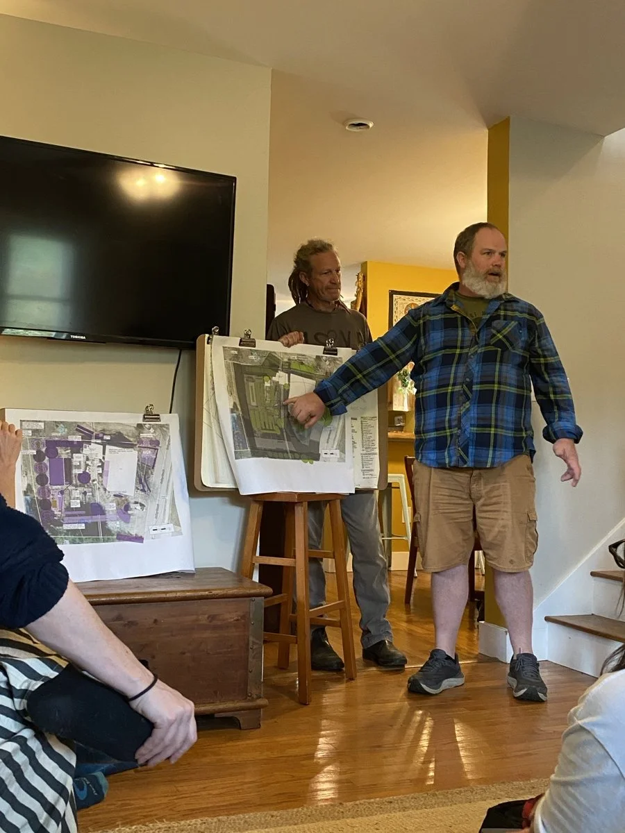 Two men presenting a project plan to an audience in a home, with one man pointing at a large map or site plan on an easel, while the other stands beside him, near a TV.