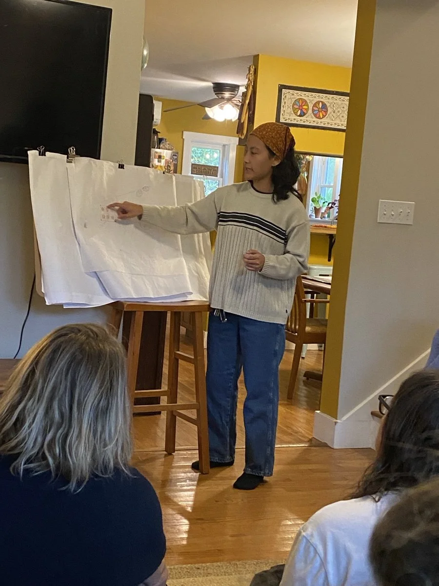 A person is giving a presentation or speech to an audience in a home setting, standing in front of a large piece of paper on an easel, with several people seated on the floor watching.