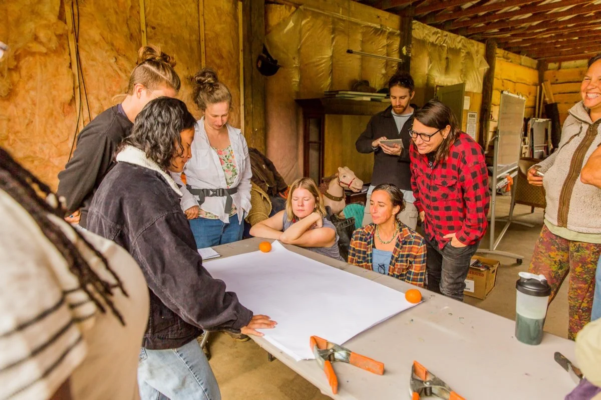 Group of people gathered around a large sheet of paper on a table in a workshop or studio, with some leaning in and discussing, while others observe.