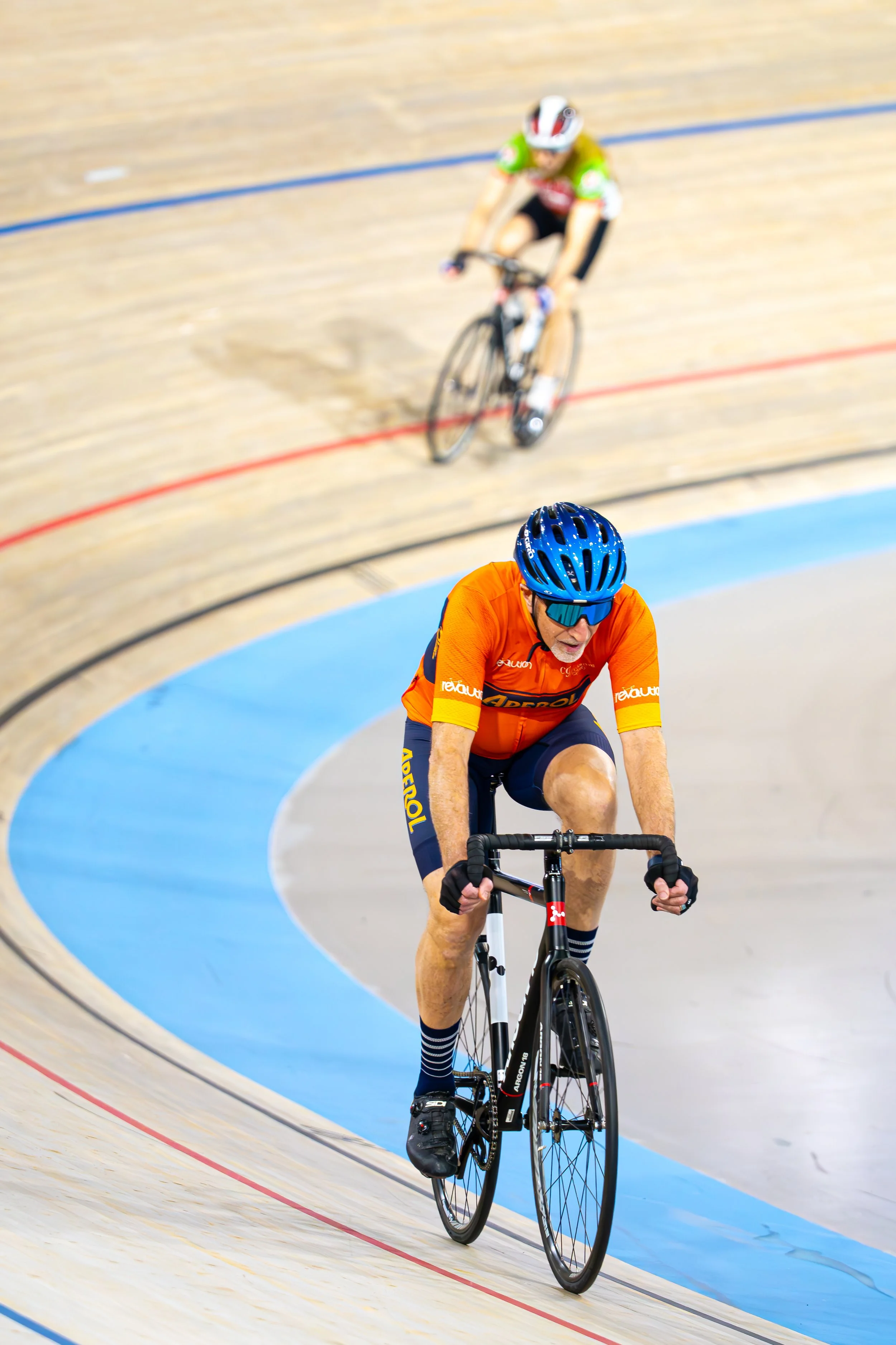 Two male cyclists racing on a velodrome, with one in orange and the other in green, all wearing cycling helmets and sunglasses.