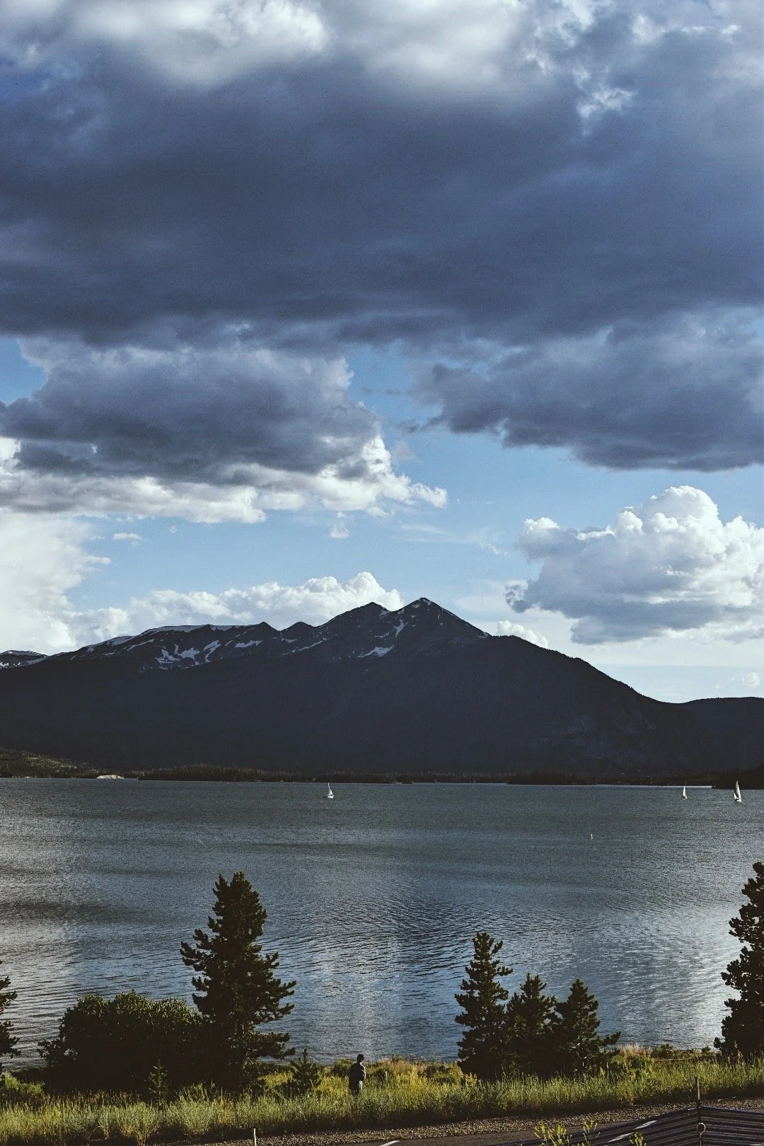 A scenic view of a mountain with patches of snow, a body of water with sailboats, and trees in the foreground under a partly cloudy sky.