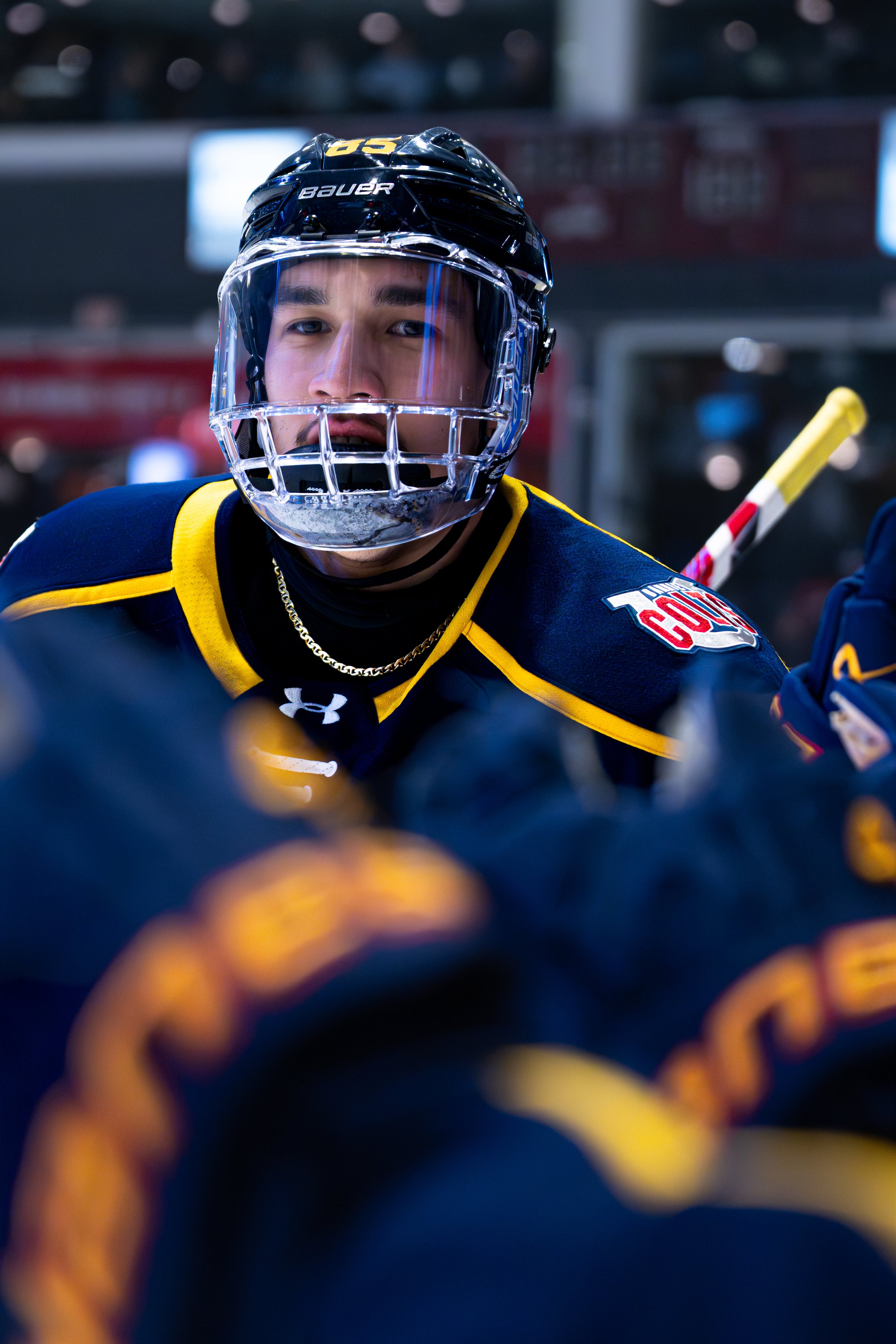 A hockey player wearing a face shield, dark jersey with yellow accents, and a black helmet with the brand 'Bauer' visible. The player appears focused, and hockey sticks and other team members are in the background.
