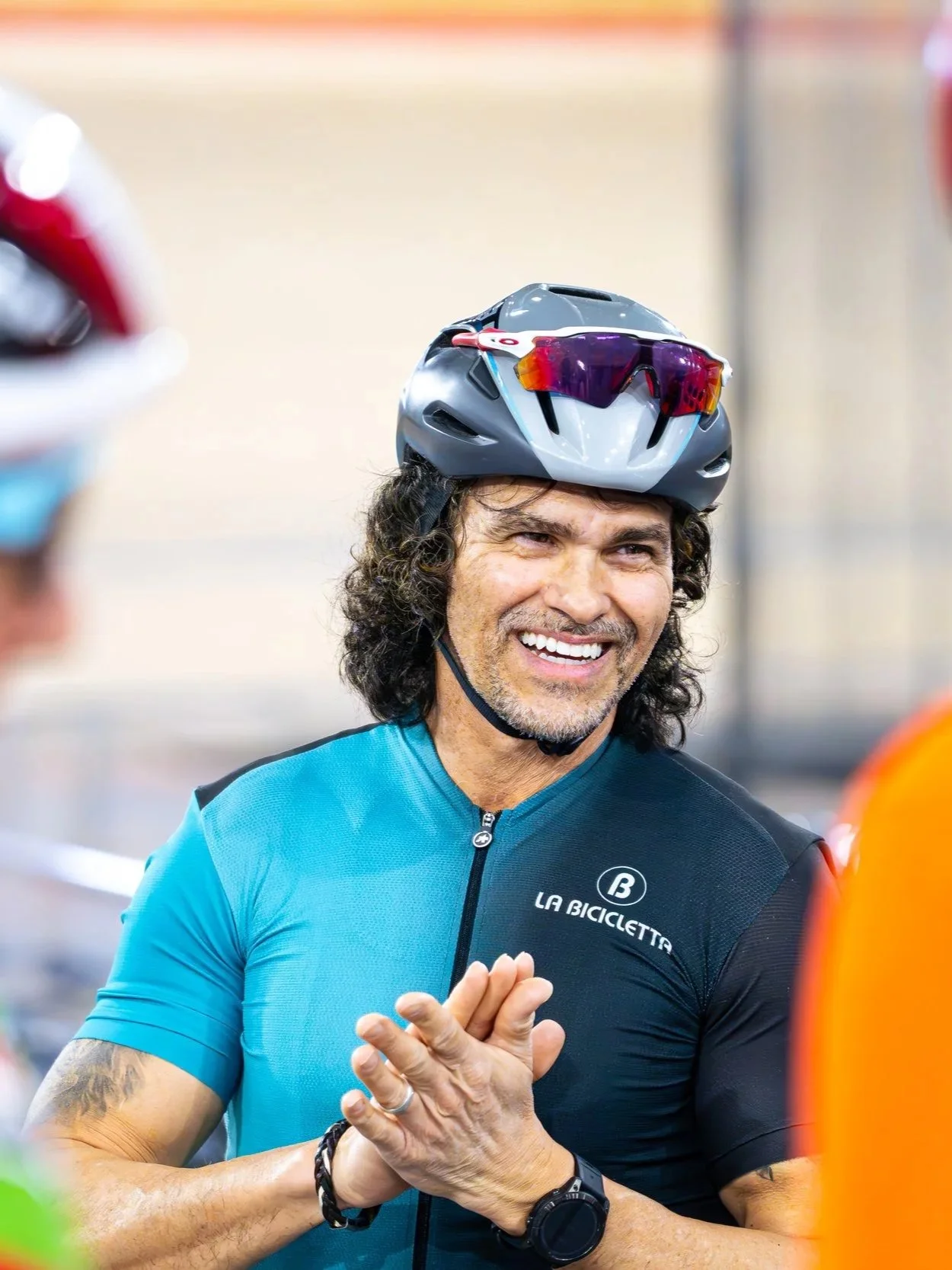 A man wearing a gray cycling helmet with sunglasses on it, a blue cycling jersey with 'La Bicicletta' logo, and a black watch, smiling and talking to others in a group.