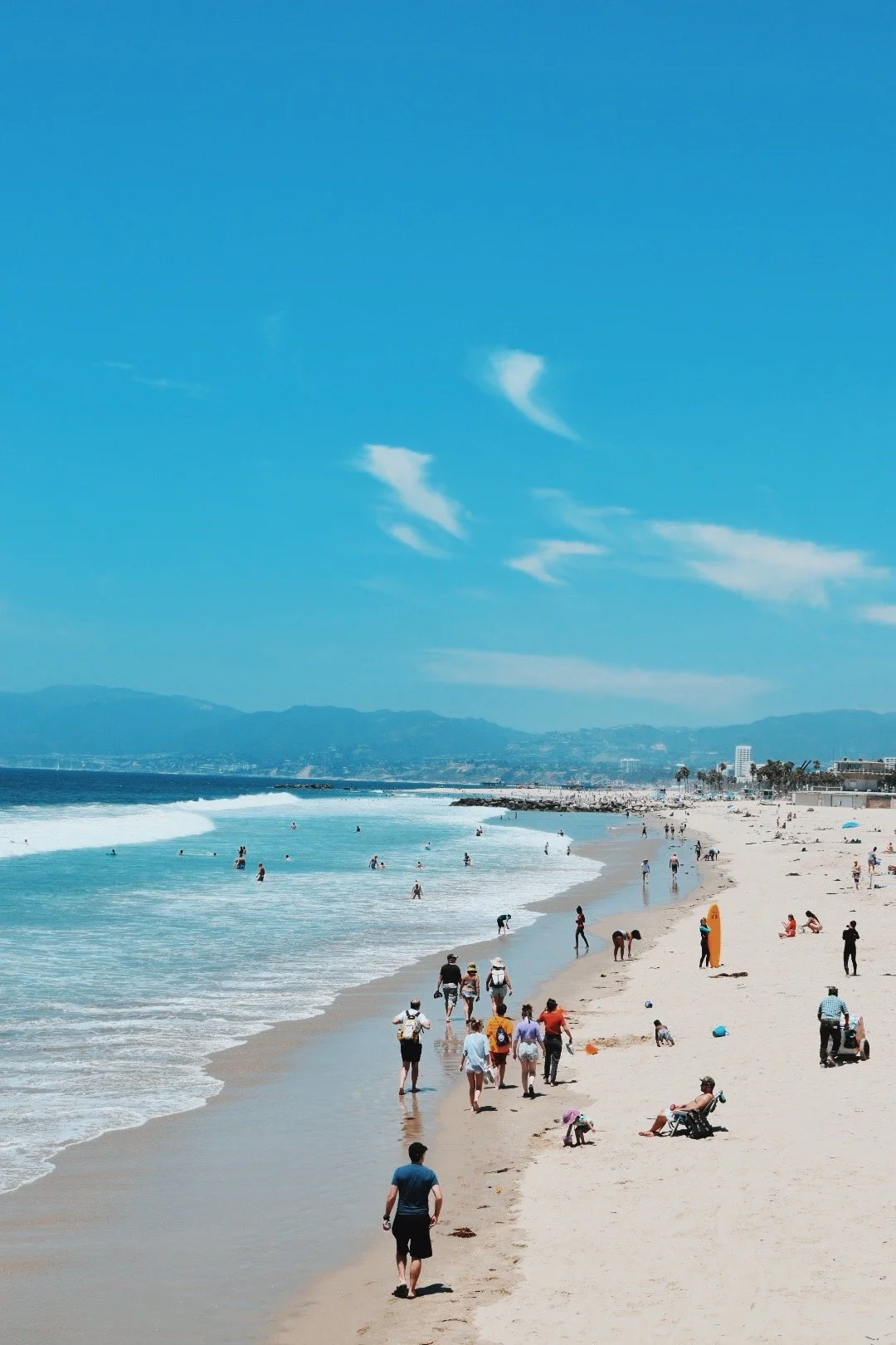 Beach scene with people walking, sitting, and swimming along the shoreline under a bright blue sky with a few clouds, distant mountains, and city buildings.