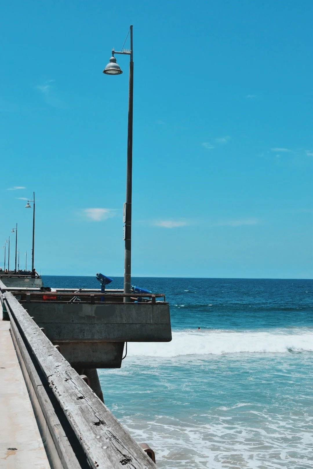 A pier with lamp posts extending into the ocean on a sunny day, with visible waves and a swimmer in the water.
