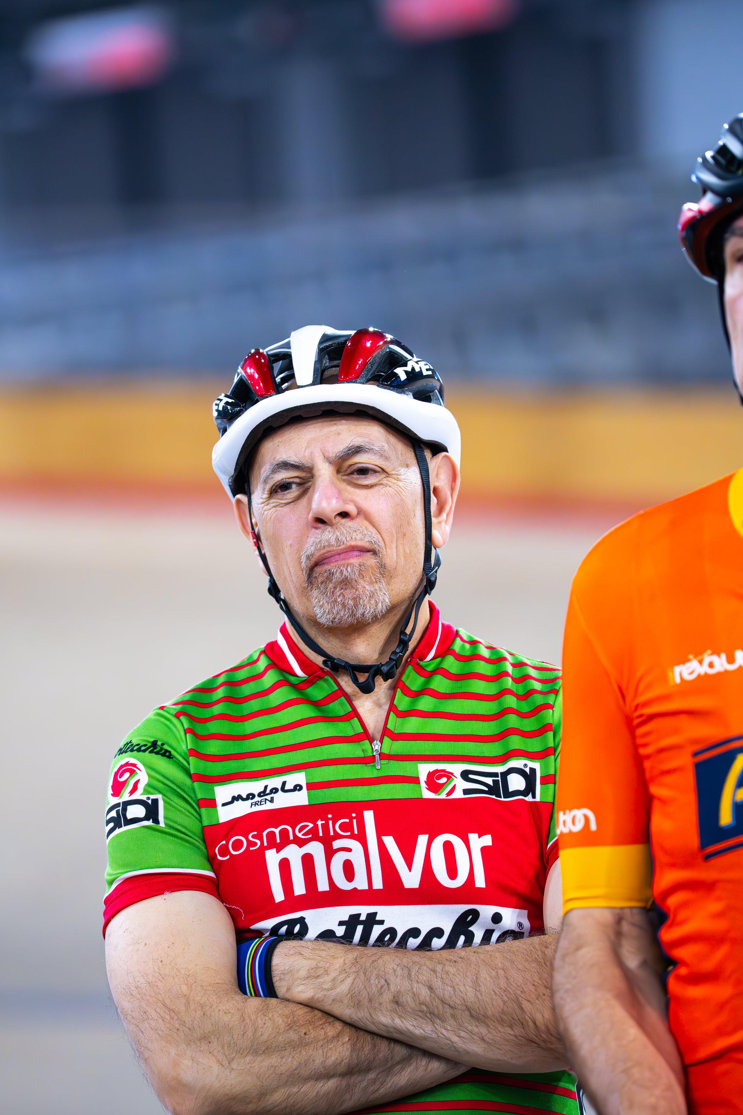 A man with gray hair and beard, wearing a green and red cycling jersey with sponsorship logos and a white helmet, standing with crossed arms and a serious expression at a cycling event.